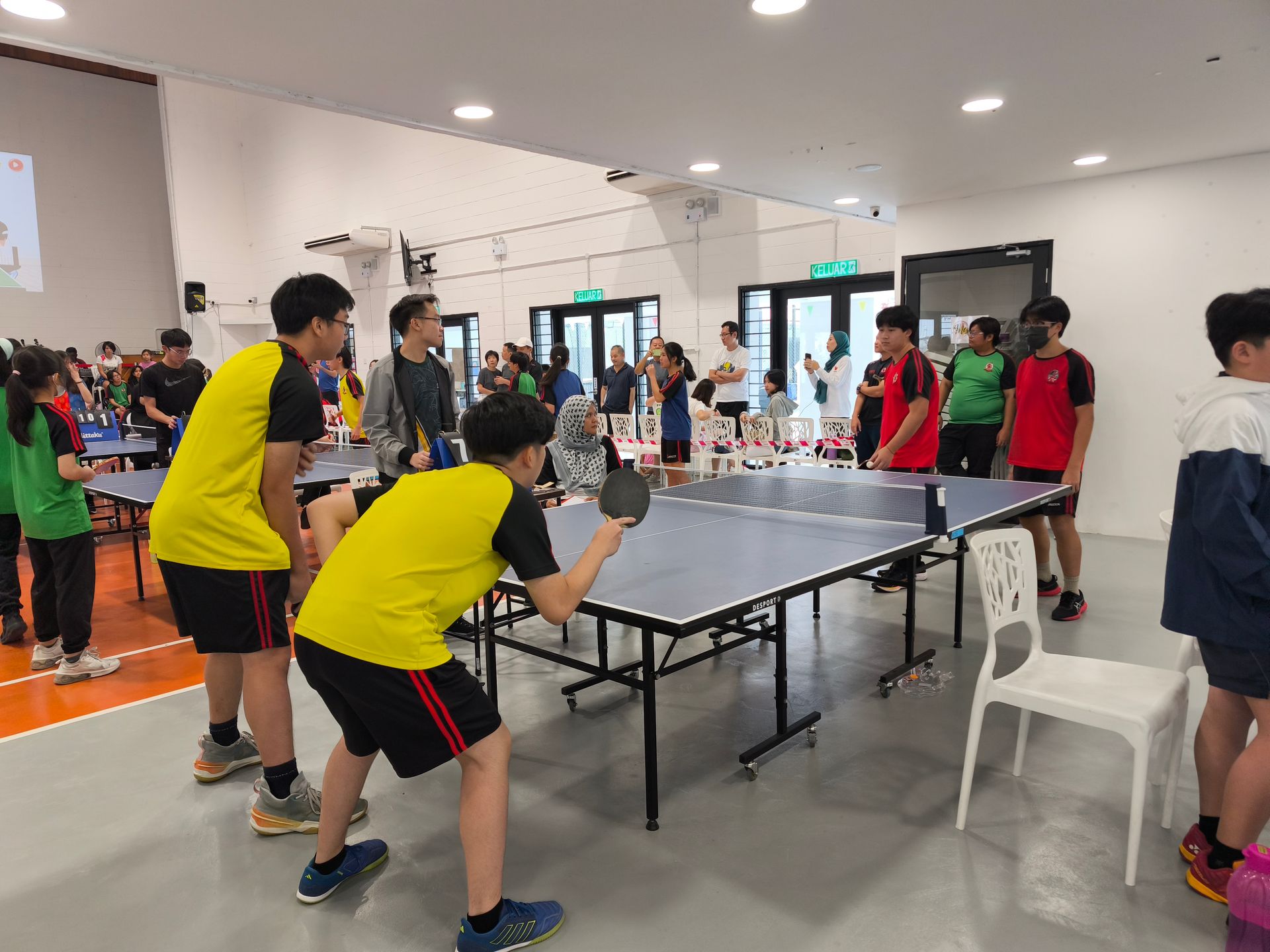 People in athletic wear play table tennis at an indoor court, with players preparing to serve across the net.