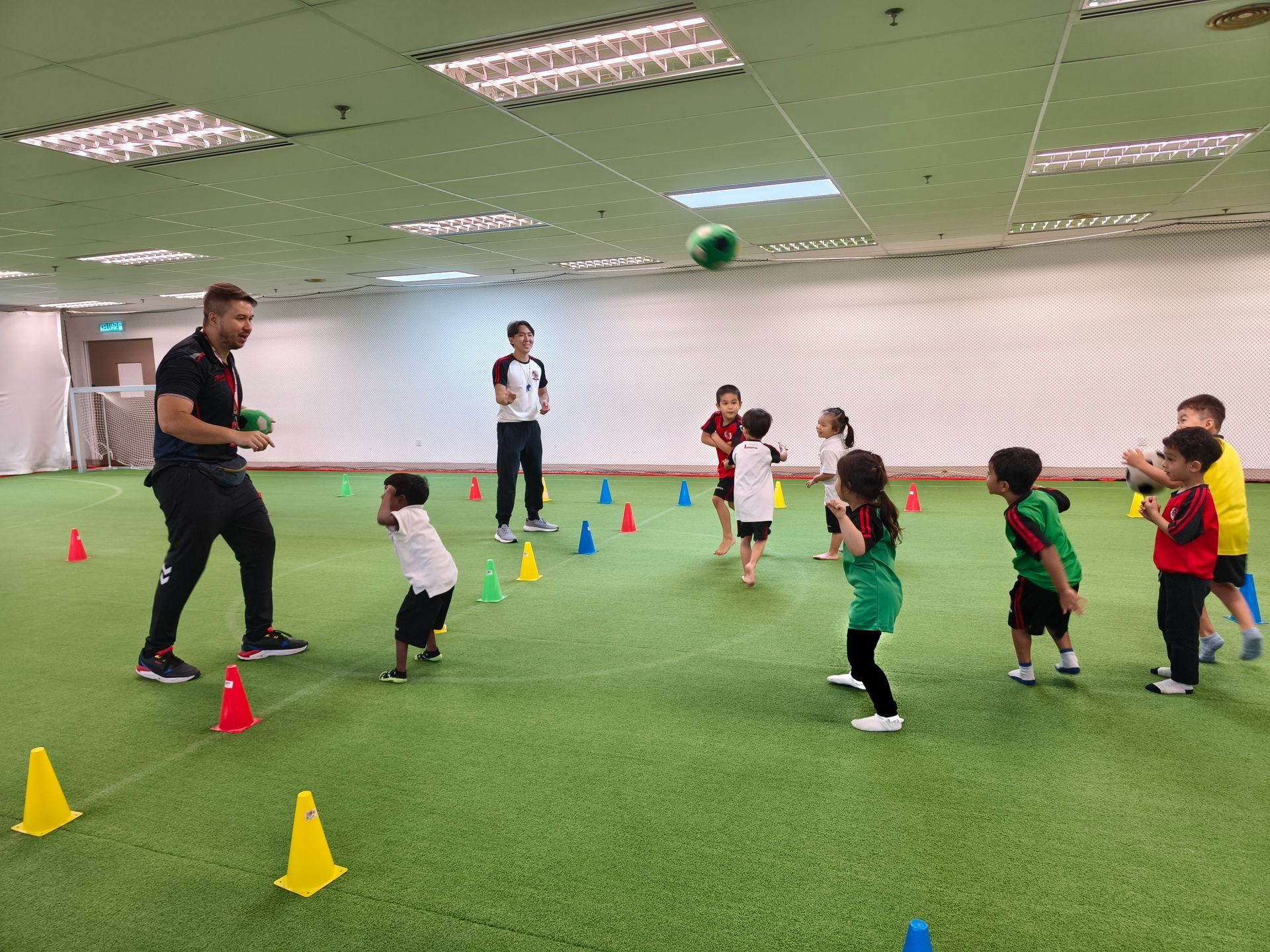A coach leading a group of young children in a sports drill with cones on an indoor green turf field as a ball is in flight.