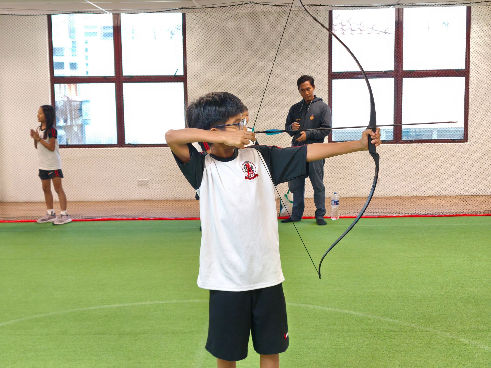 A person practices archery with a recurve bow indoors on a green field, while others stand in the background.