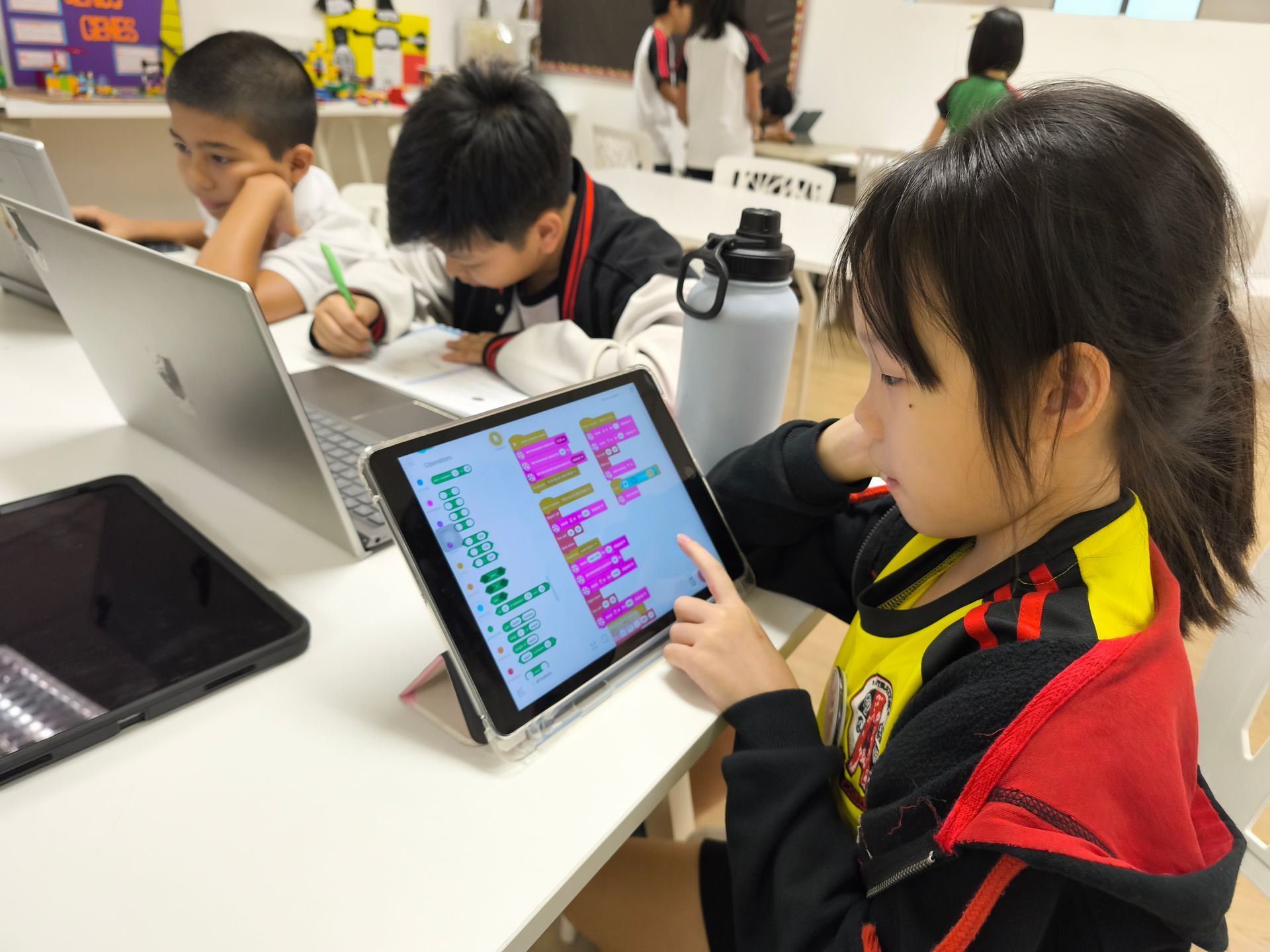 Students sit at a table in a classroom, focused on coding block-based programs on their individual laptops and tablets.