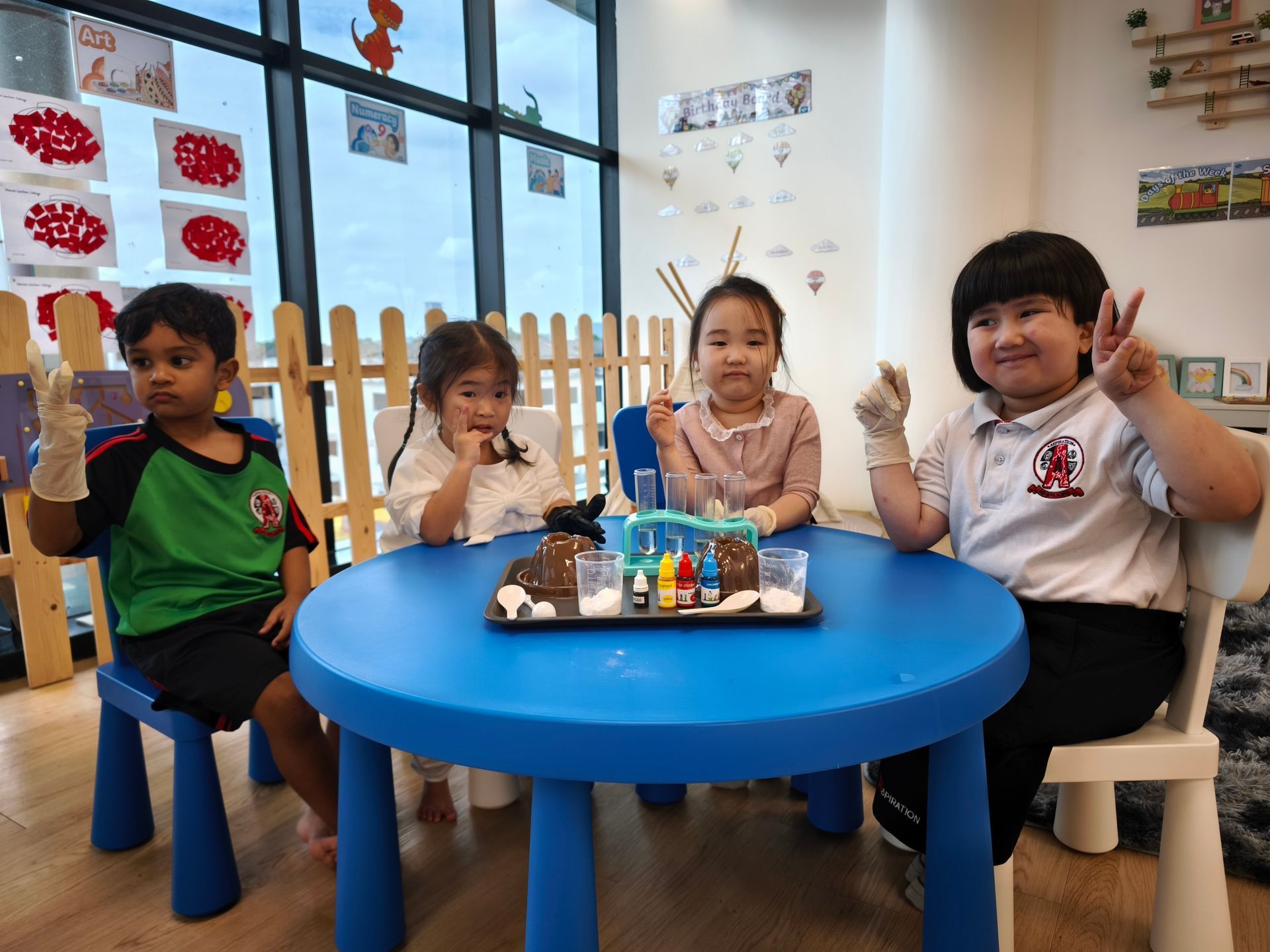 Four children in a brightly lit classroom sit at a round blue table with art supplies, smiling and wearing gloves.