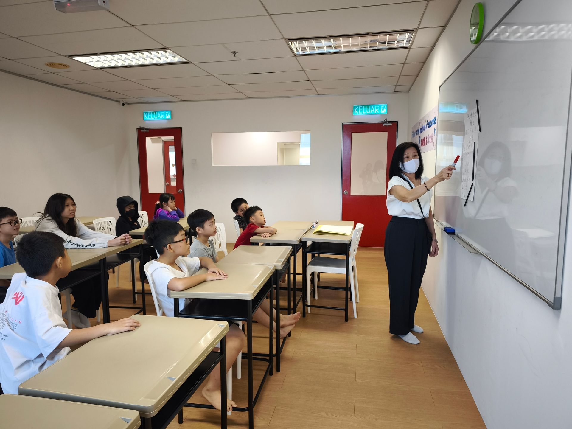 A teacher writes on a whiteboard in a classroom as a group of students sit at desks and watch.