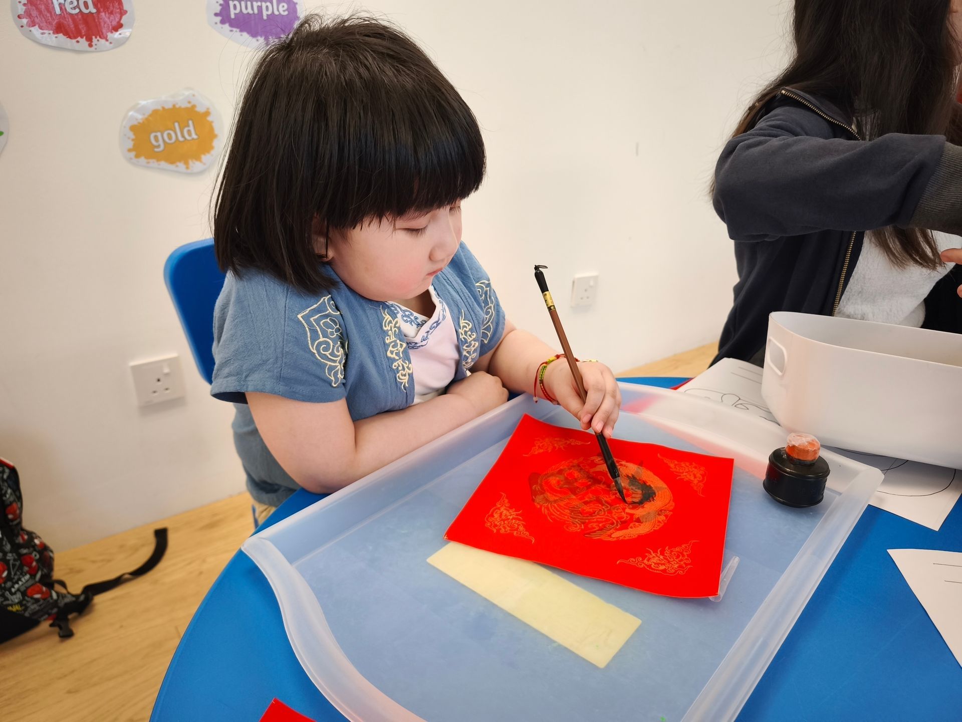 A person uses a brush and ink to paint on a red square of paper placed on a tray at a table.