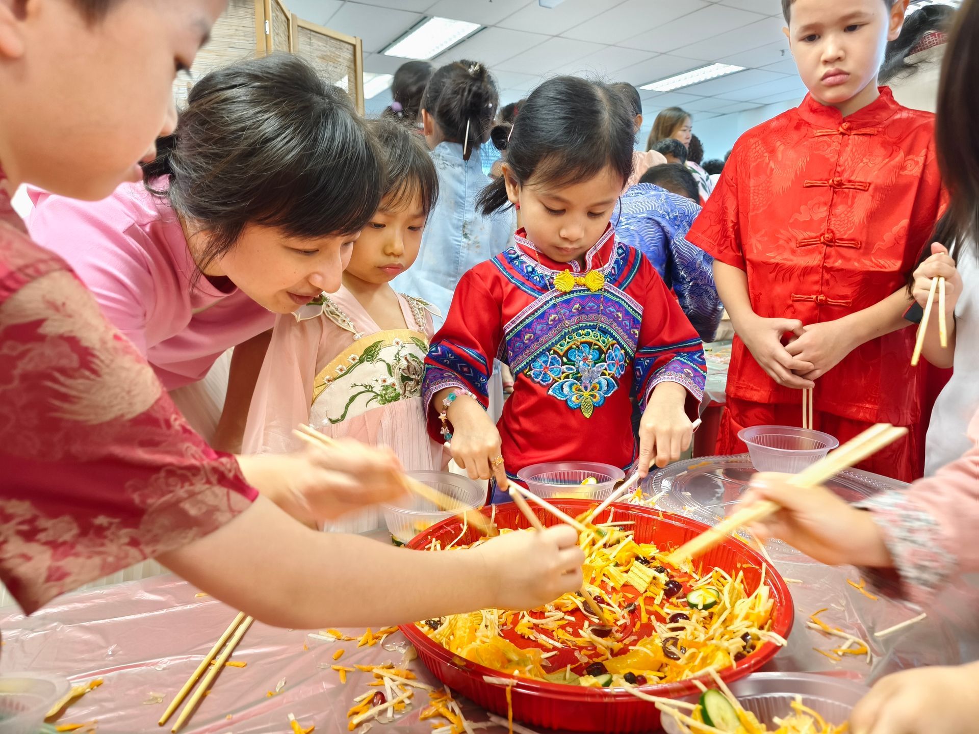 A group of people wearing traditional clothing stand around a large red bowl, using chopsticks to toss a colorful salad.