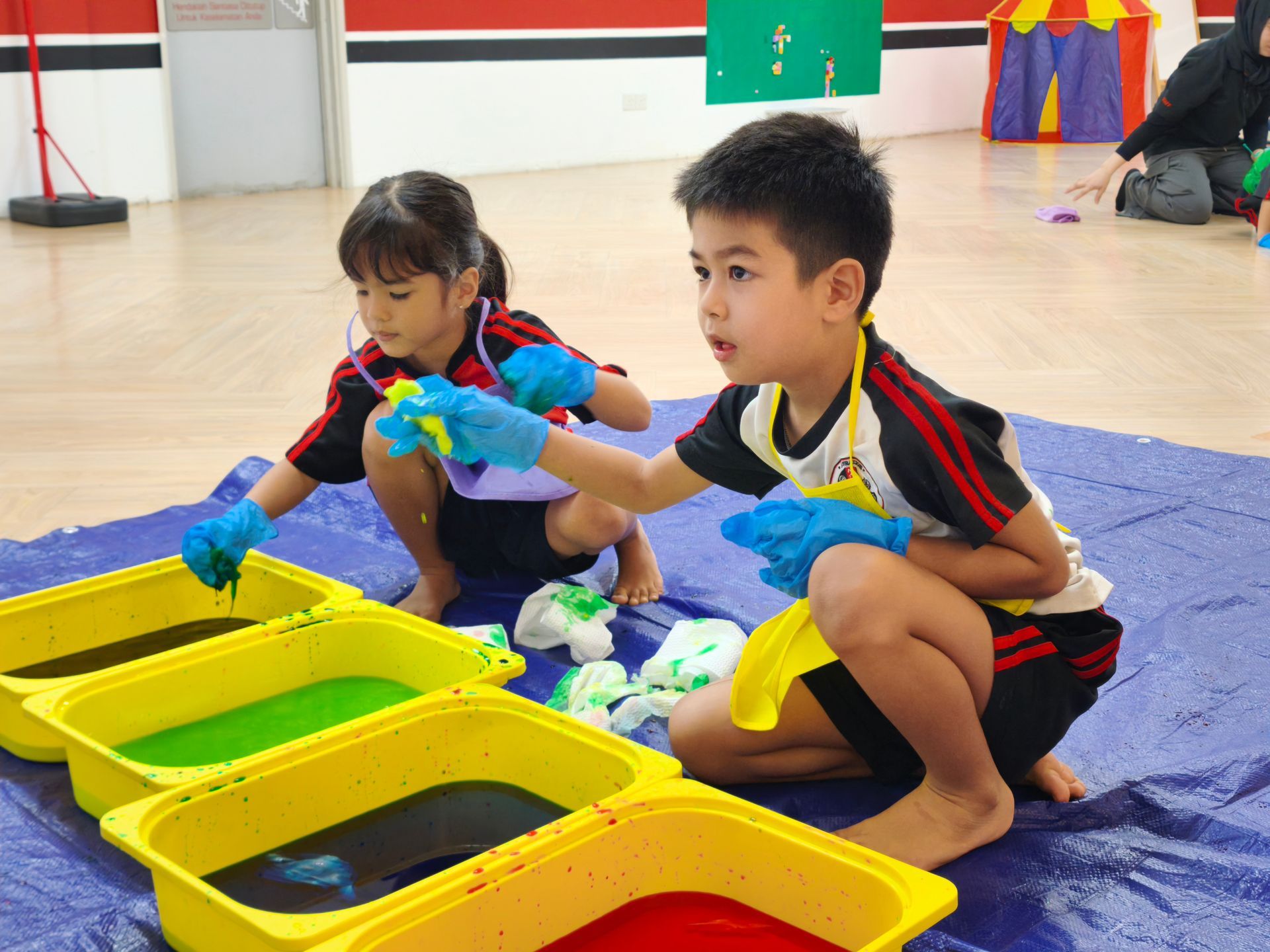 Two children wearing blue gloves squat on a blue mat, dipping sponges into four yellow bins filled with colored liquid.