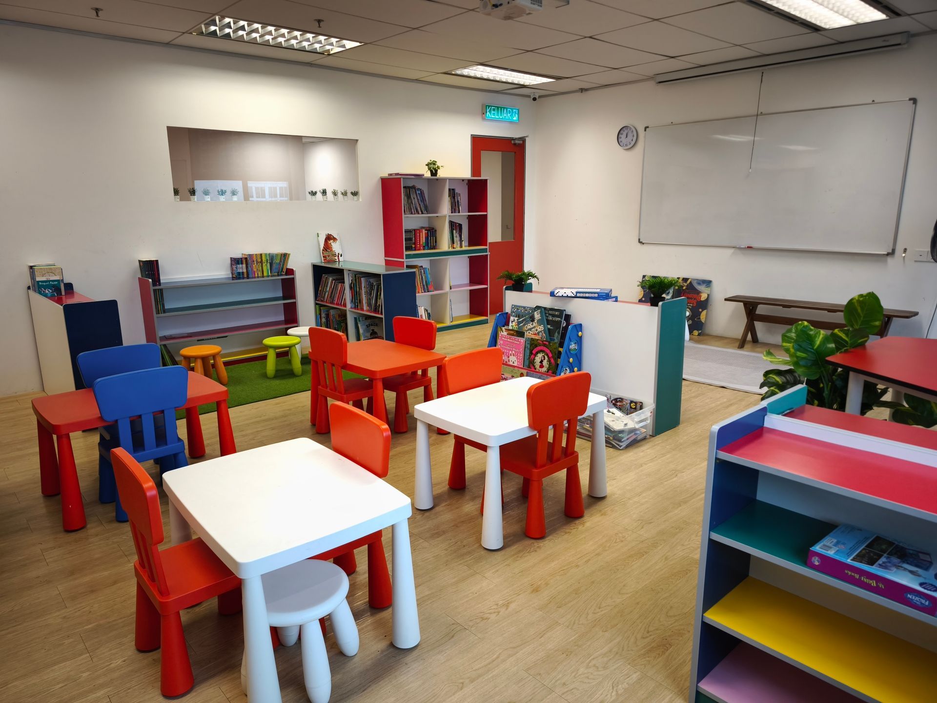 A brightly lit children's classroom with colorful small tables, chairs, and bookshelves on a wood-look floor.
