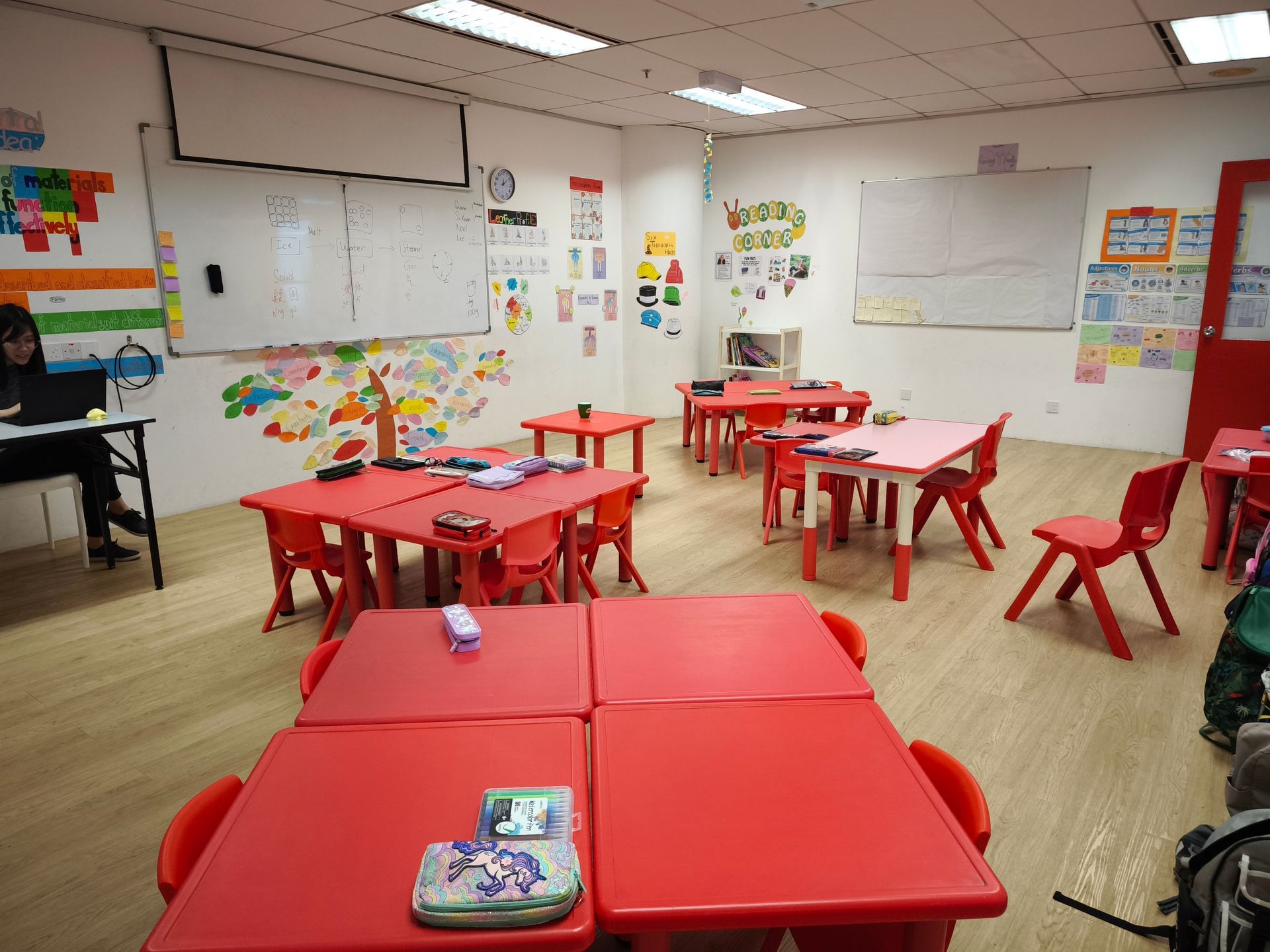Brightly lit classroom with red tables, chairs, and colorful wall decorations, featuring a teacher sitting at a desk.