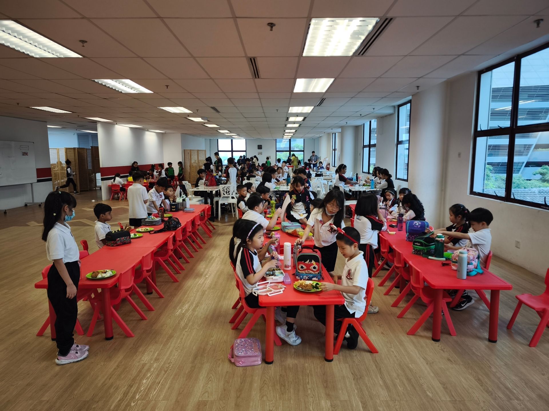 Students sitting at red tables in a brightly lit room, eating during what appears to be a school meal or event.