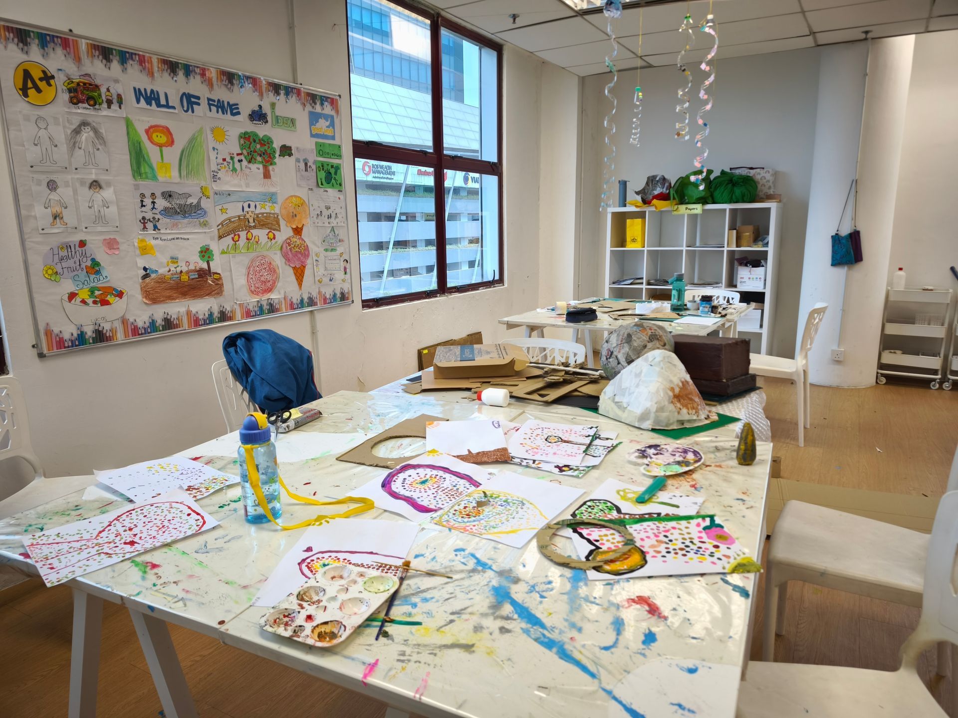 An art classroom features a paint-splattered table covered in drawings, a water bottle, and a display board of artwork.