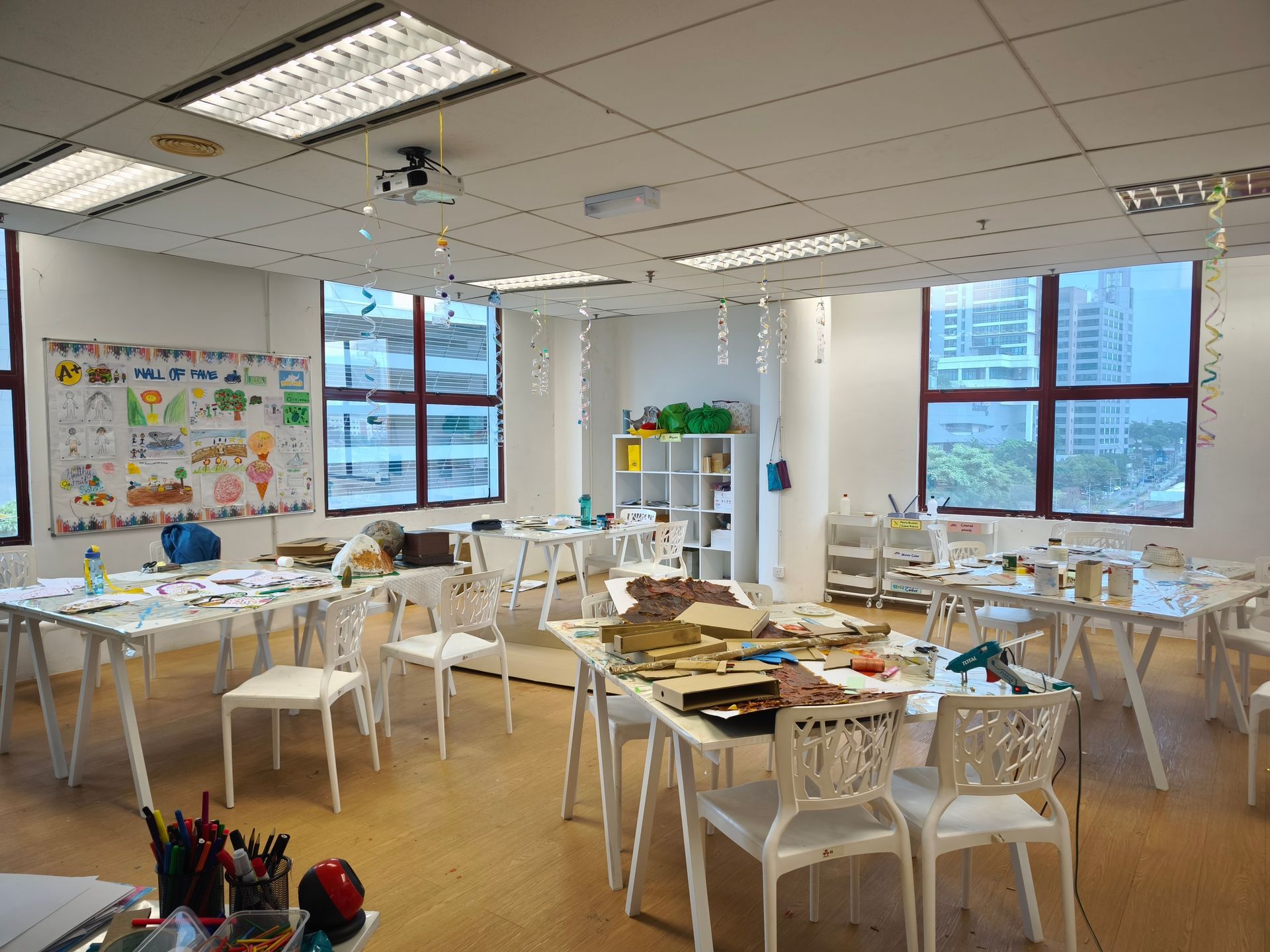 A brightly lit classroom with white tables, chairs, art supplies, and decorative hanging ornaments.