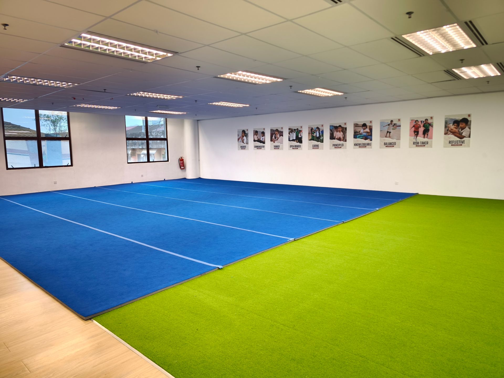 An indoor gym features a large blue mat area next to a green turf surface, with posters on the white wall.