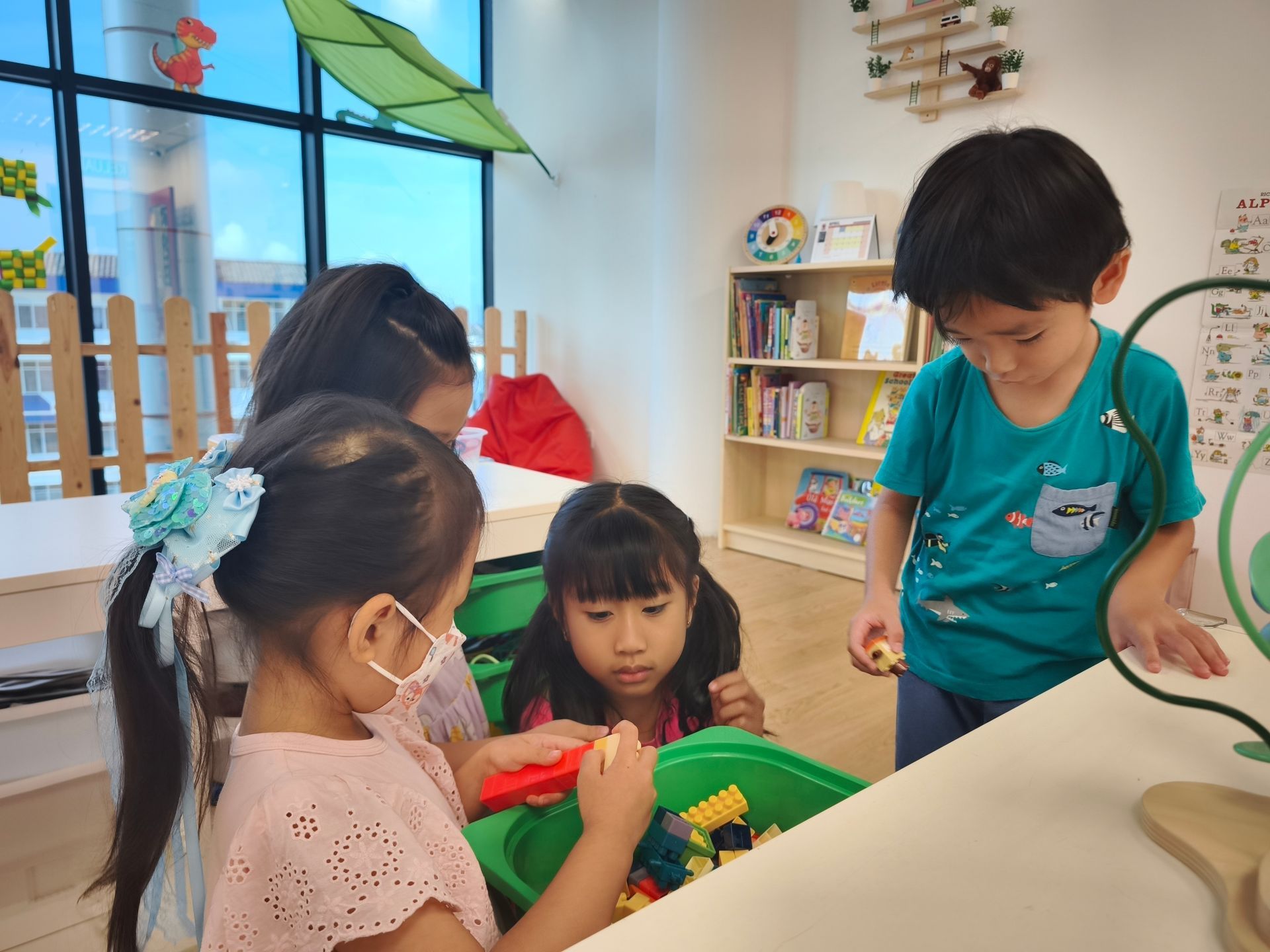 Children sit around a green bin, playing with colorful building blocks in a bright, organized classroom.