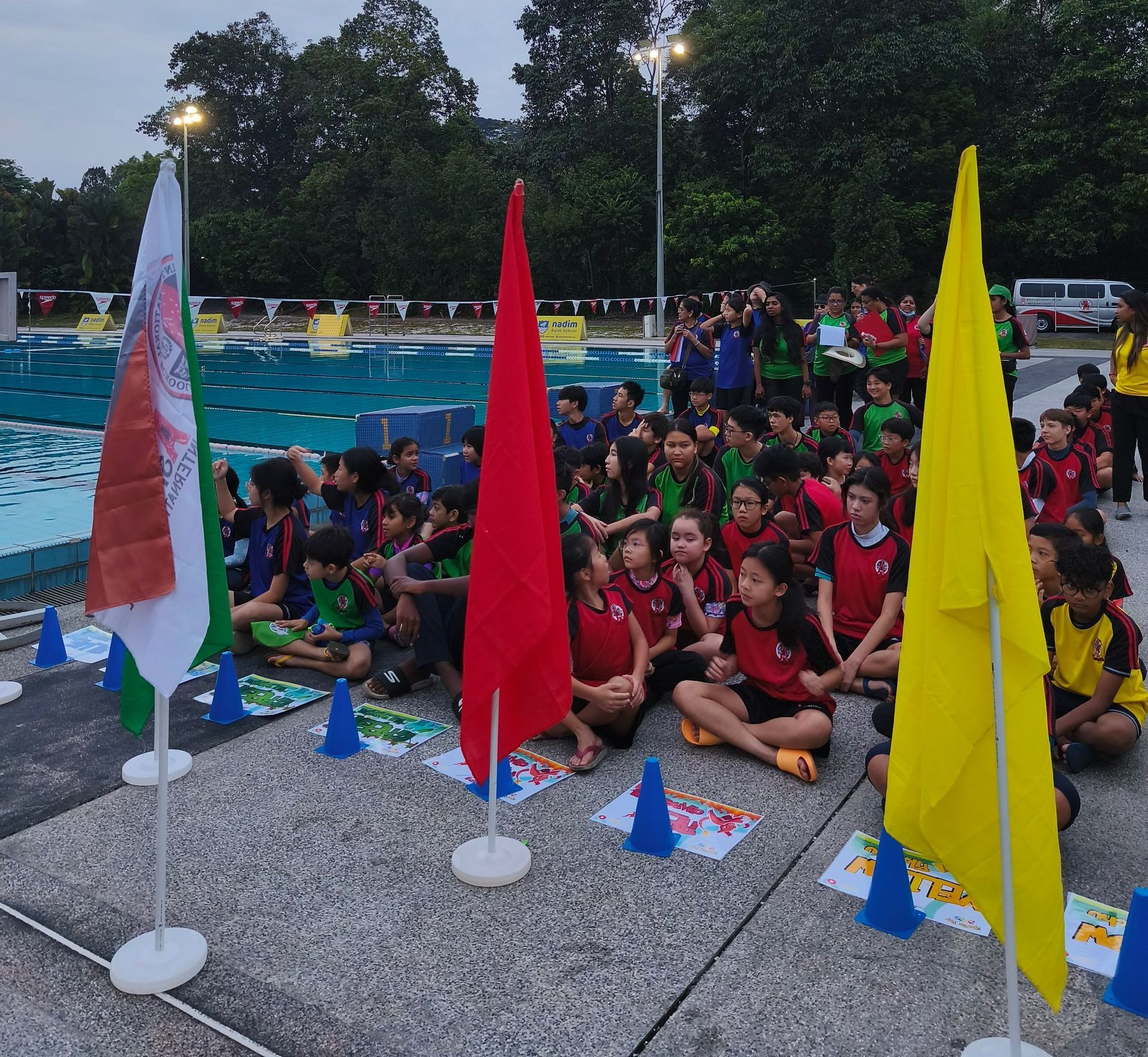 A group of students in team uniforms sits on a pool deck near white, red, and yellow flags during an outdoor swim event.