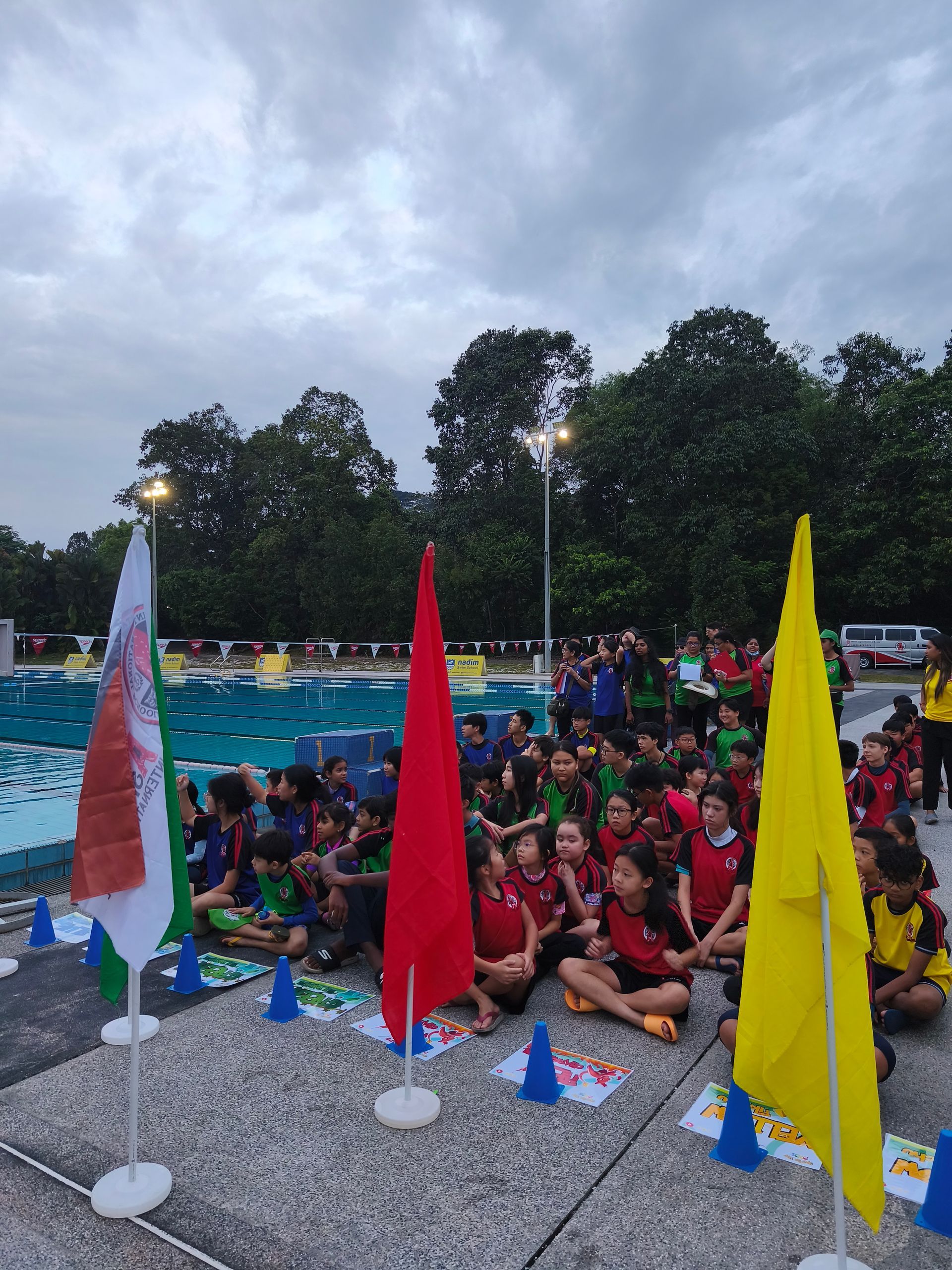 A crowd of people sits on a pool deck behind three flags—white, red, and yellow—facing a swimming pool.