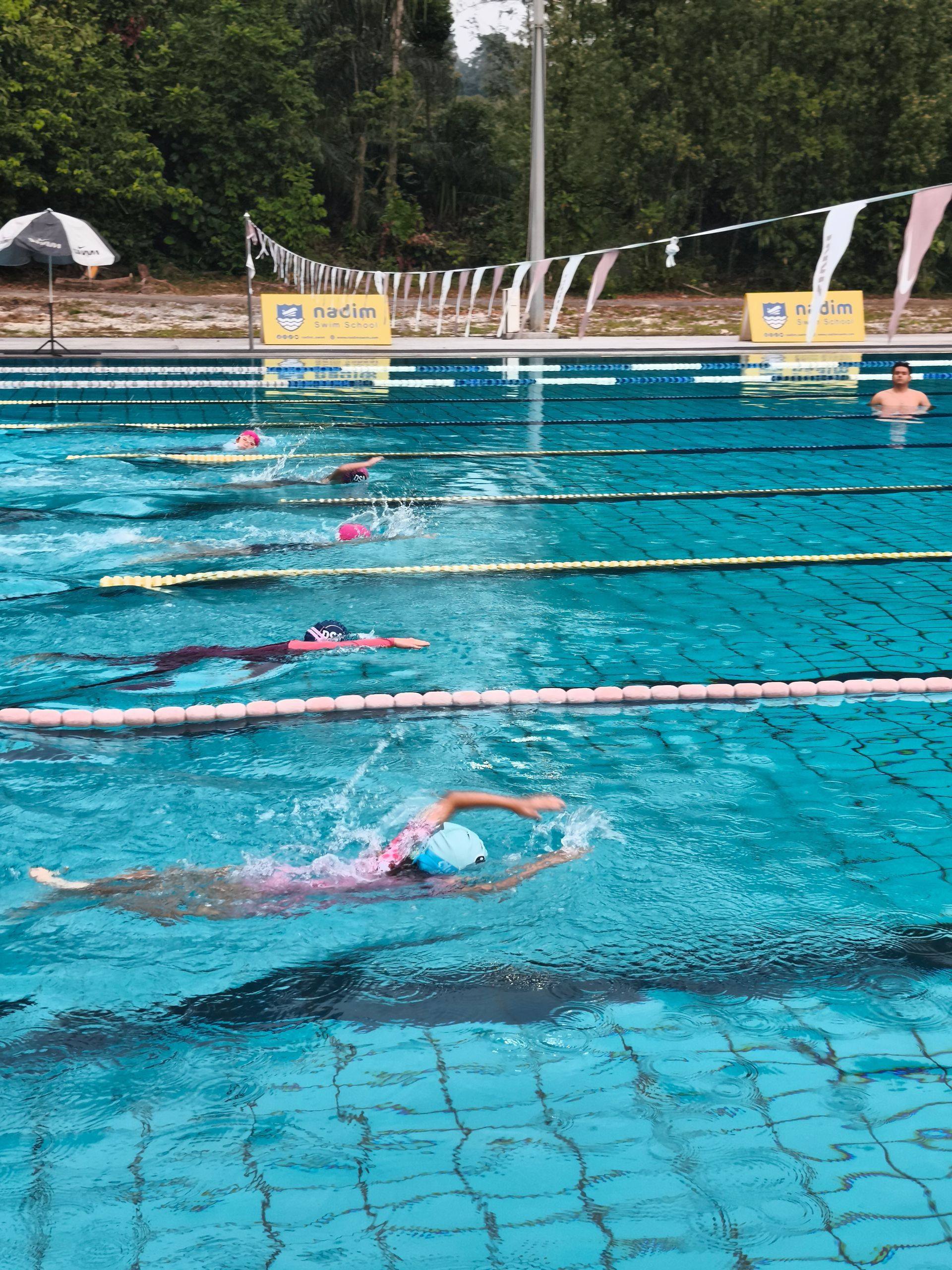 Swimmers in a training pool performing laps, with lane dividers, flags, and a person watching from the water.