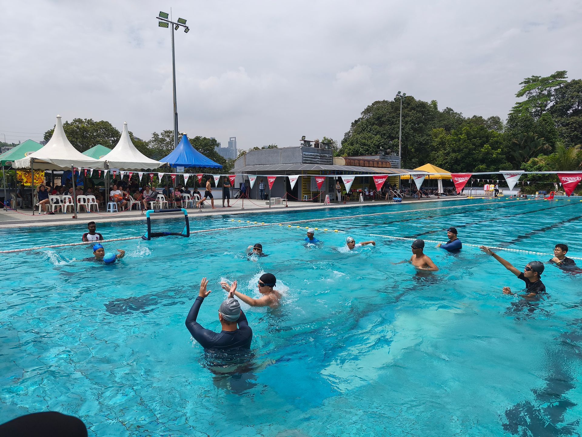 People in swimsuits participate in a group water exercise class in an outdoor pool under cloudy skies.