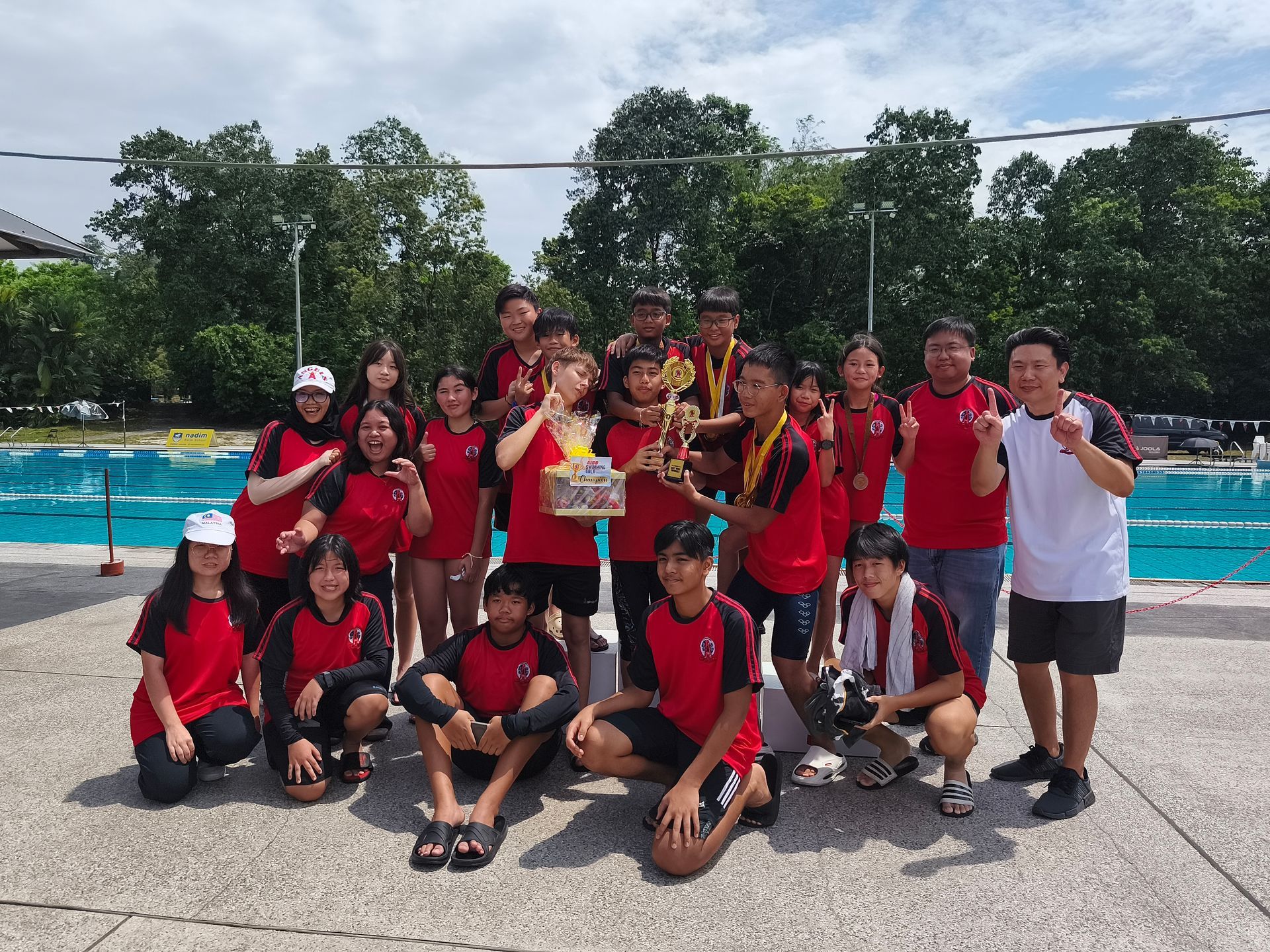 A group in red and black uniforms poses with a trophy poolside under a sunny sky.