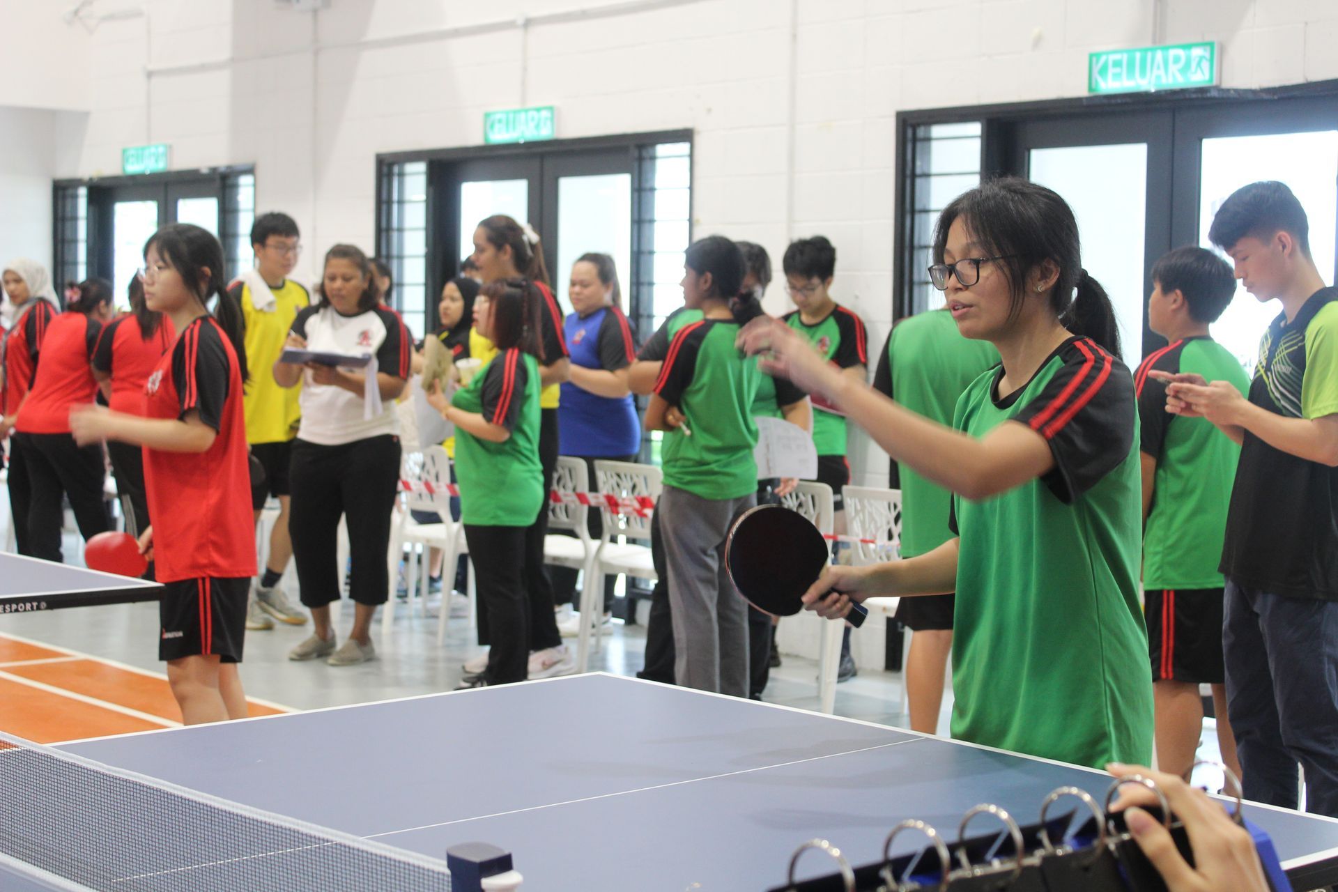 People in athletic attire gather at a table tennis tournament in an indoor facility.