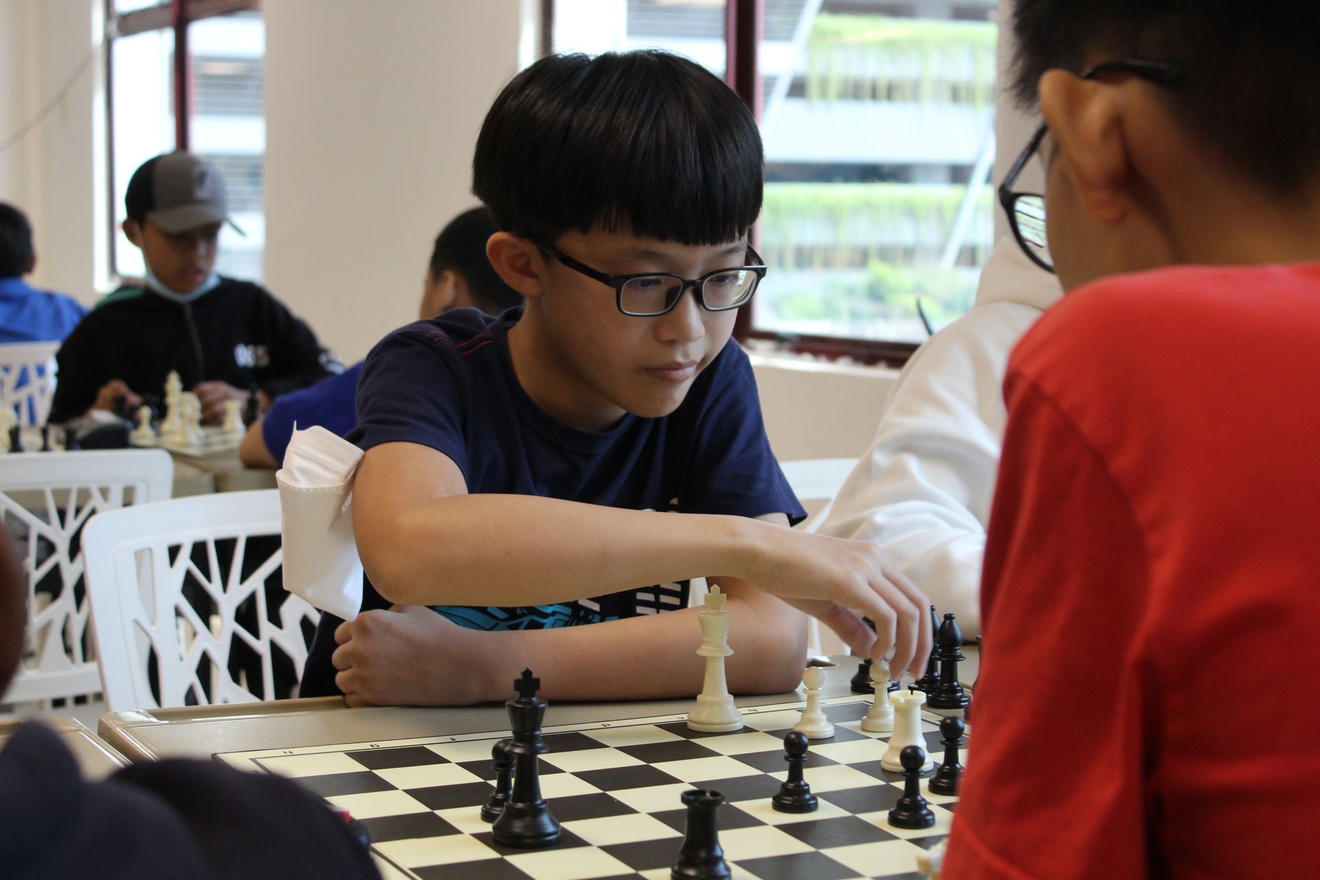 Two chess players focus on their game in a bright room, with one moving a white piece on the board.