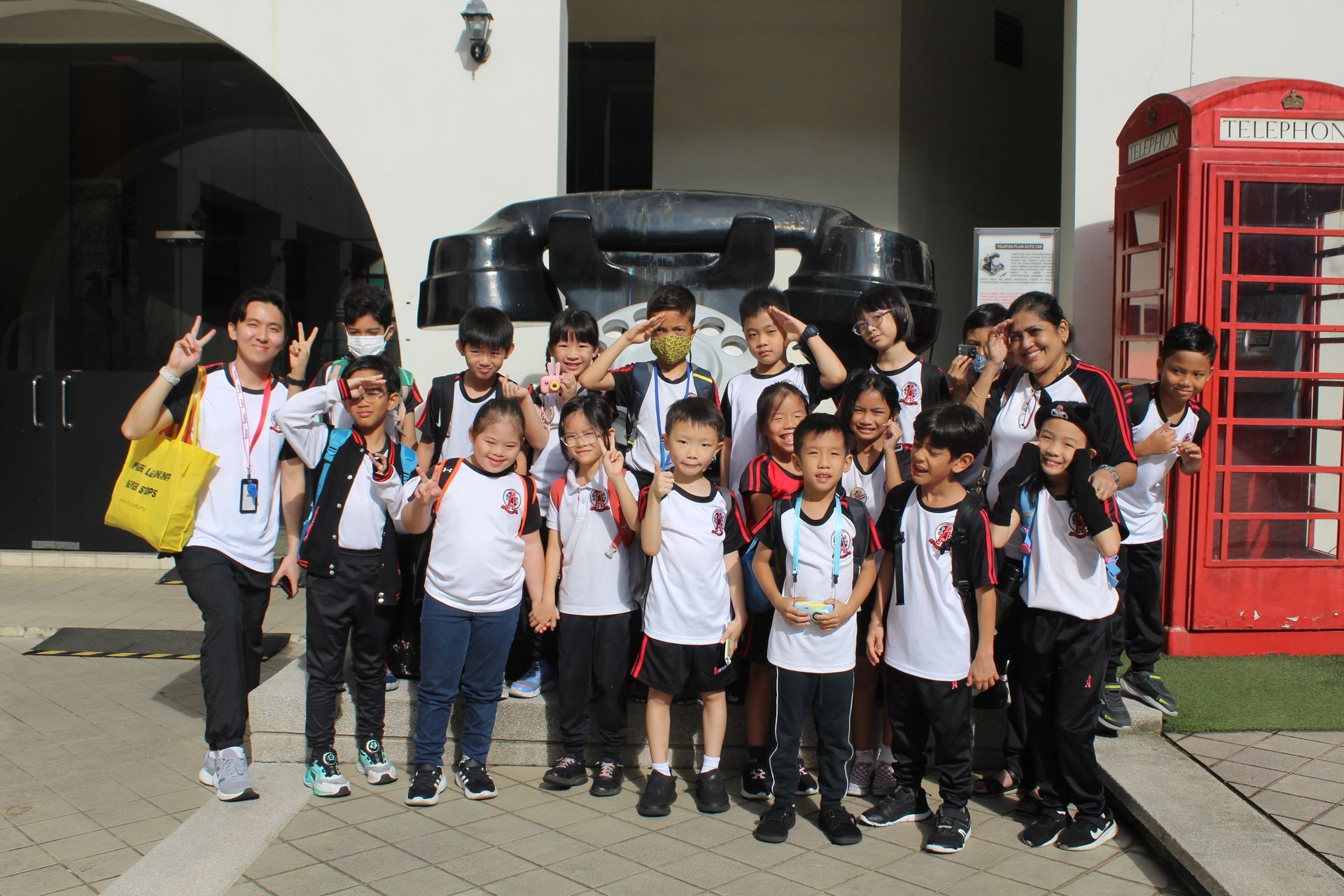 A group of children and adults pose together outdoors in front of a giant telephone sculpture and a red phone booth.