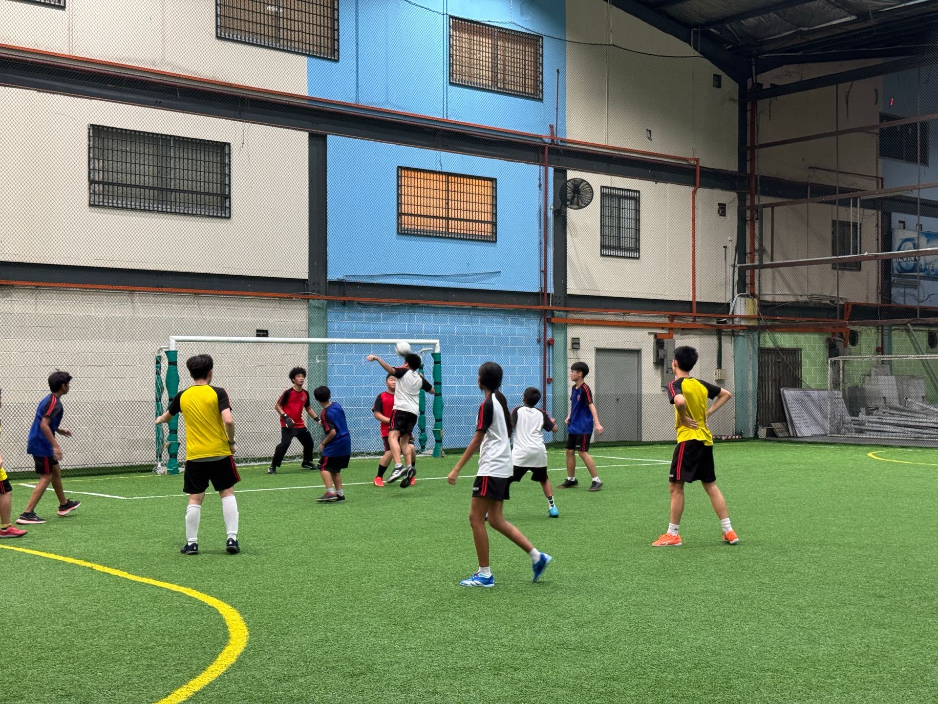 A group of people in athletic wear playing a soccer game on an indoor green turf field with a goal net.