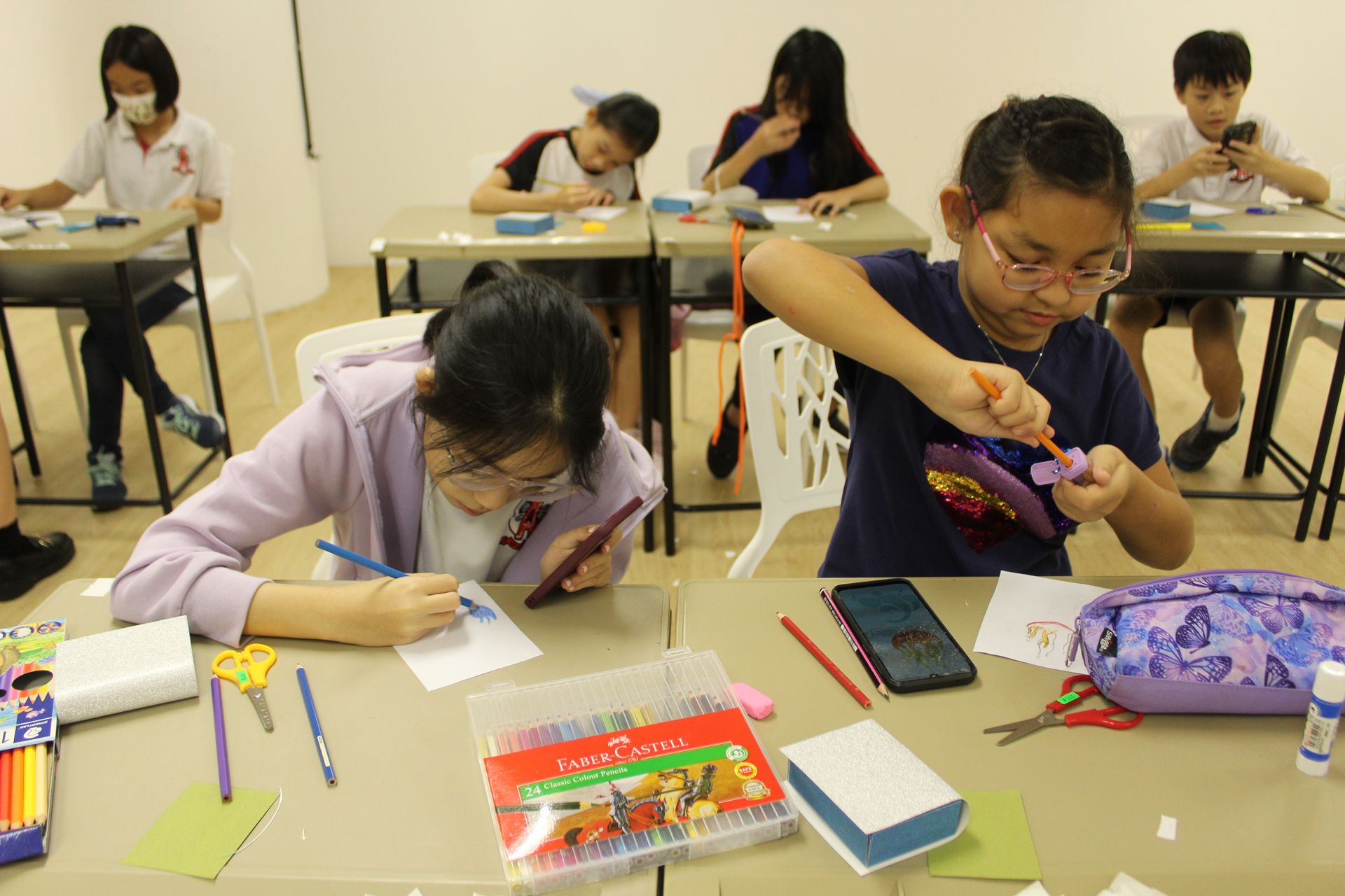 Students sit at desks in a classroom, focused on individual craft projects, coloring, and using tools with art supplies.