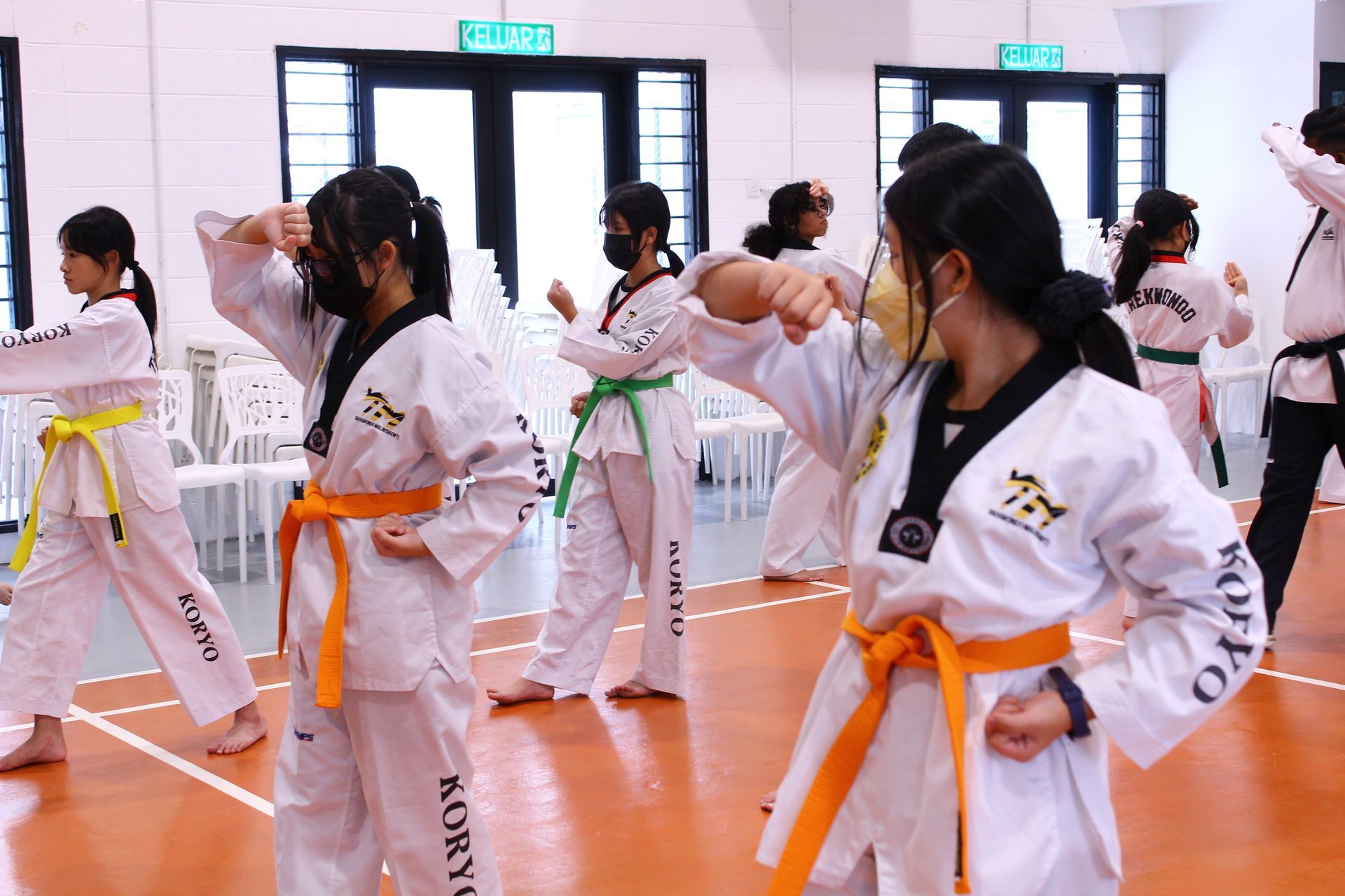 A group of taekwondo students in white uniforms with colored belts practicing martial arts movements in a gym.