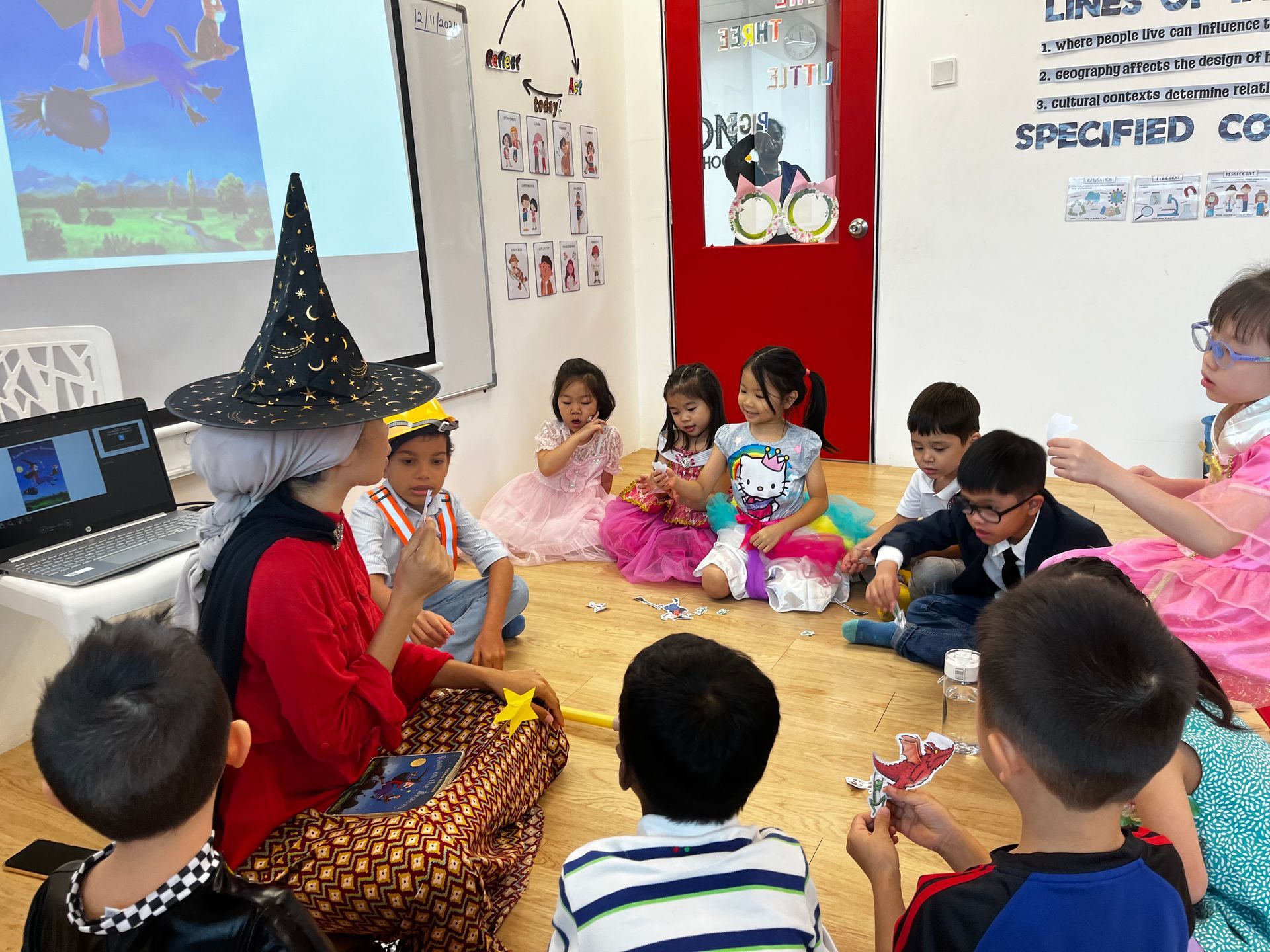 A person in a witch hat leads a group of children sitting on the floor in a classroom for a storytelling activity.