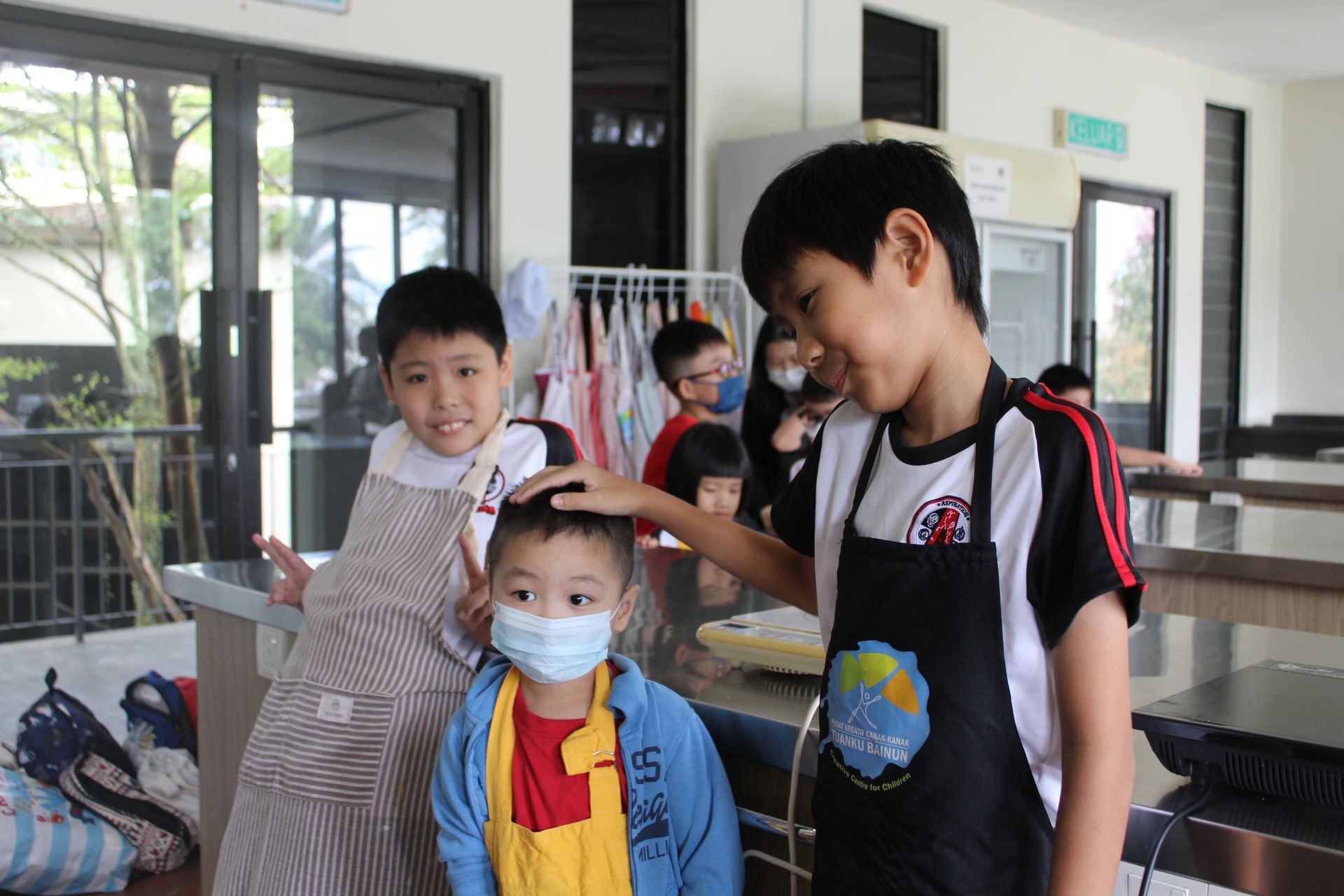 Three children in a kitchen-like setting; one wears a face mask, another wears a black apron, and a third wears a smock.