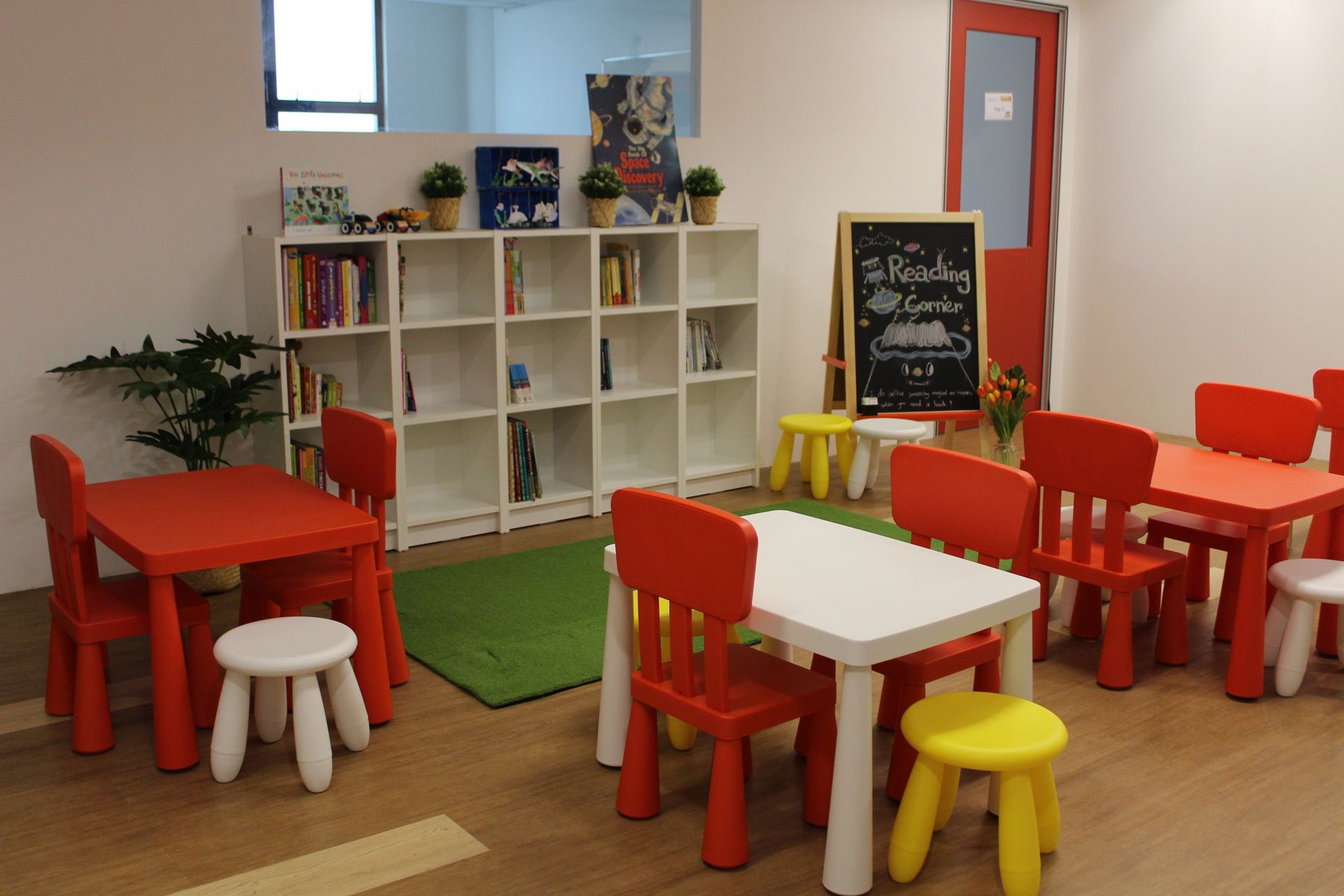 A bright playroom with small, colorful tables and chairs, a white bookshelf, and a chalkboard sign reading 