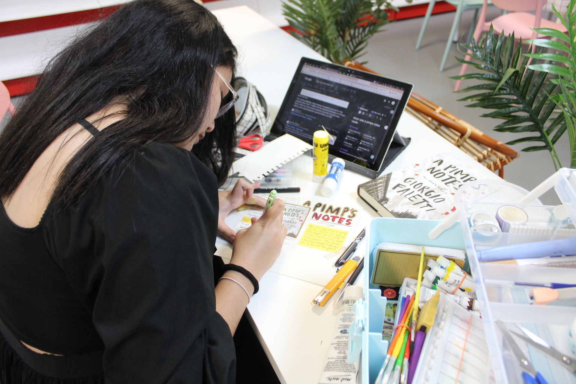 A person sits at a desk, focused on writing in a notebook near a laptop, craft supplies, and a plant in a bright room.