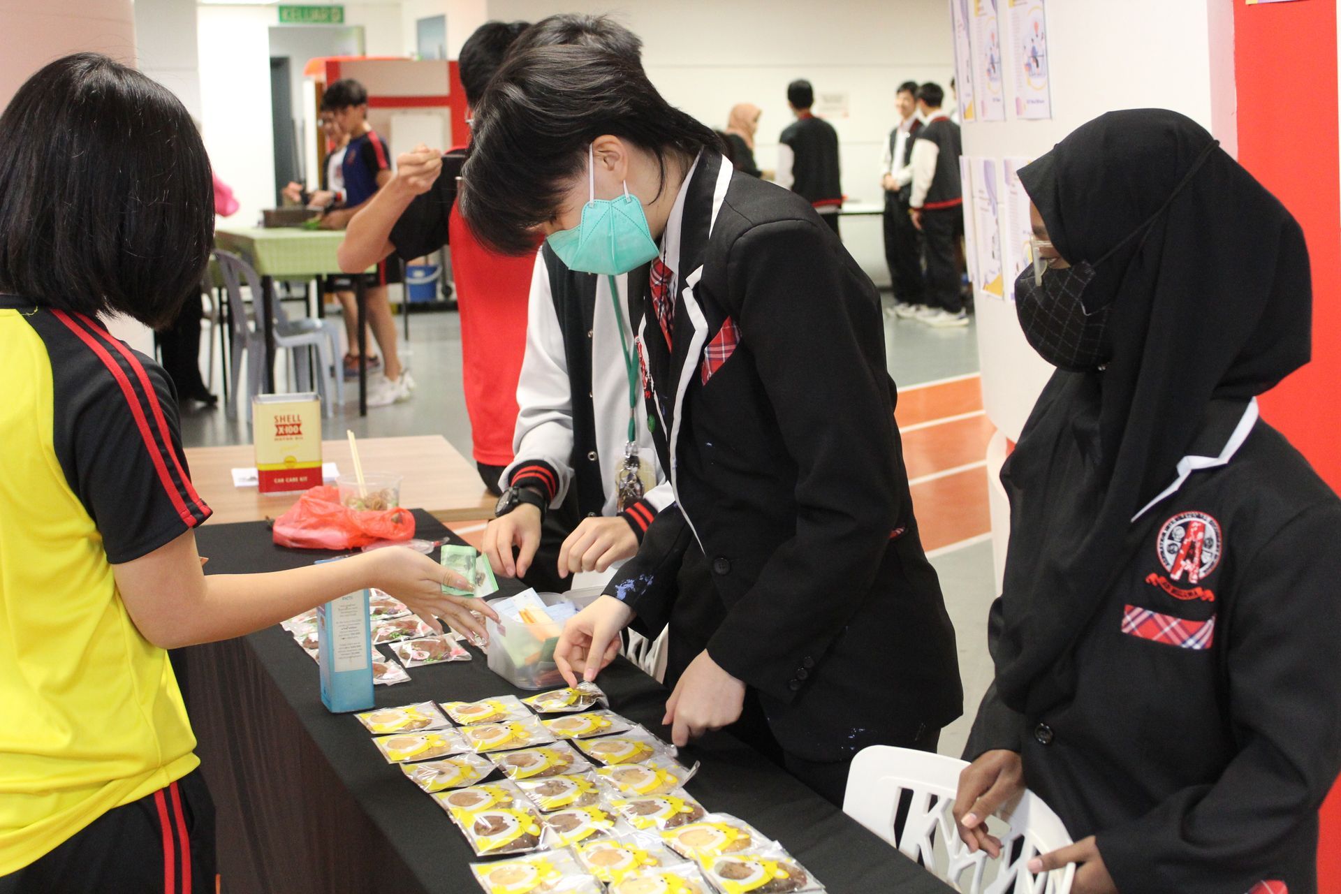 Students in school uniforms distribute wrapped snacks to a peer at a table in a brightly lit indoor venue.