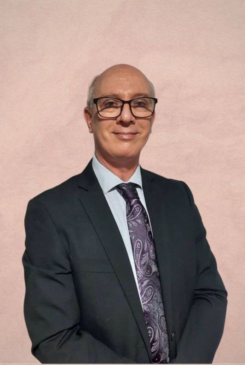 A person wearing glasses, a dark suit, a light shirt, and a patterned purple tie against a light pink wall.