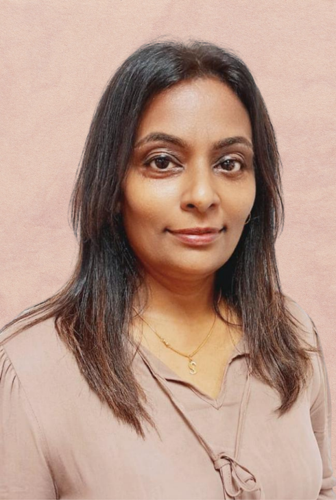 A person with long dark hair wearing a light brown top and a gold necklace, posed against a solid, textured pink background.