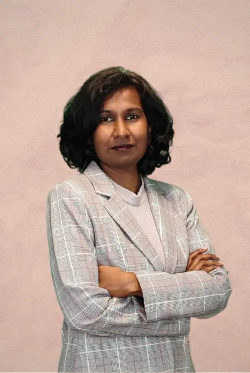 A person with dark hair styled in a bob, wearing a grey plaid blazer over a light shirt, standing with arms crossed.