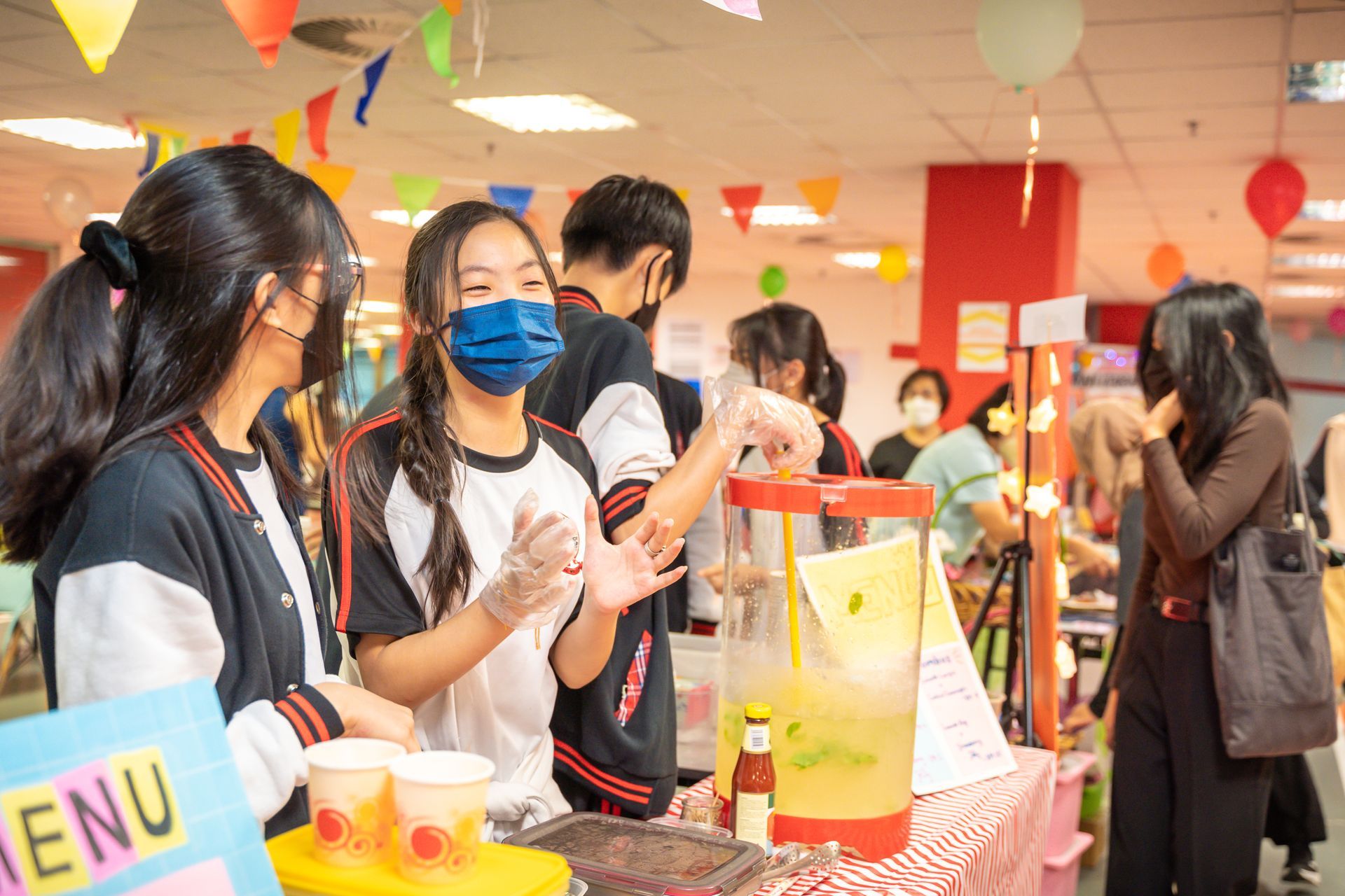 Students behind a food stall at an indoor event with decorations, serving drinks from a large dispenser.