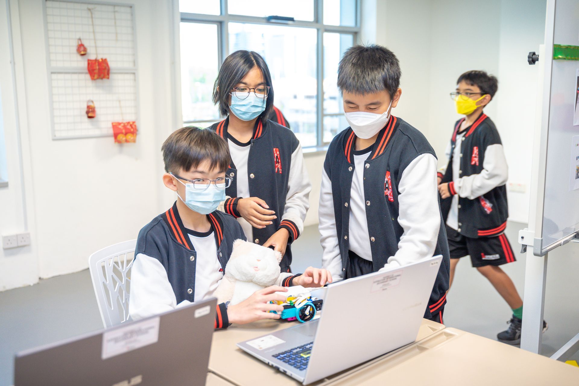 Students in school uniforms wearing face masks work together on a laptop and small robotic project in a classroom.