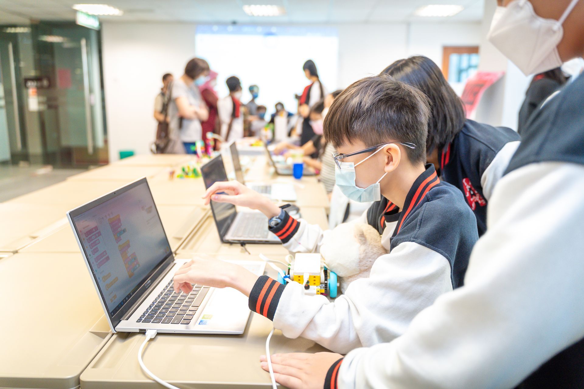 Students wearing face masks work together on laptops and robotic projects at a desk in a classroom setting.