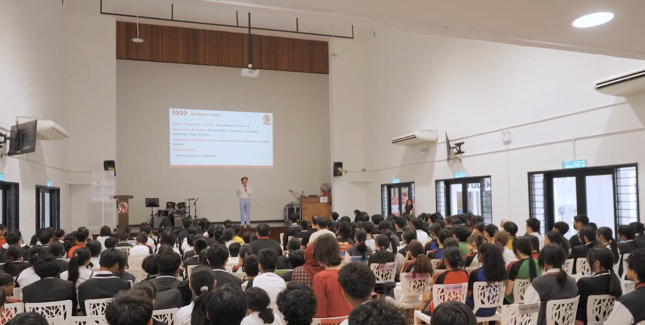 A speaker addresses a large audience gathered in a spacious hall for a presentation with a projection screen.