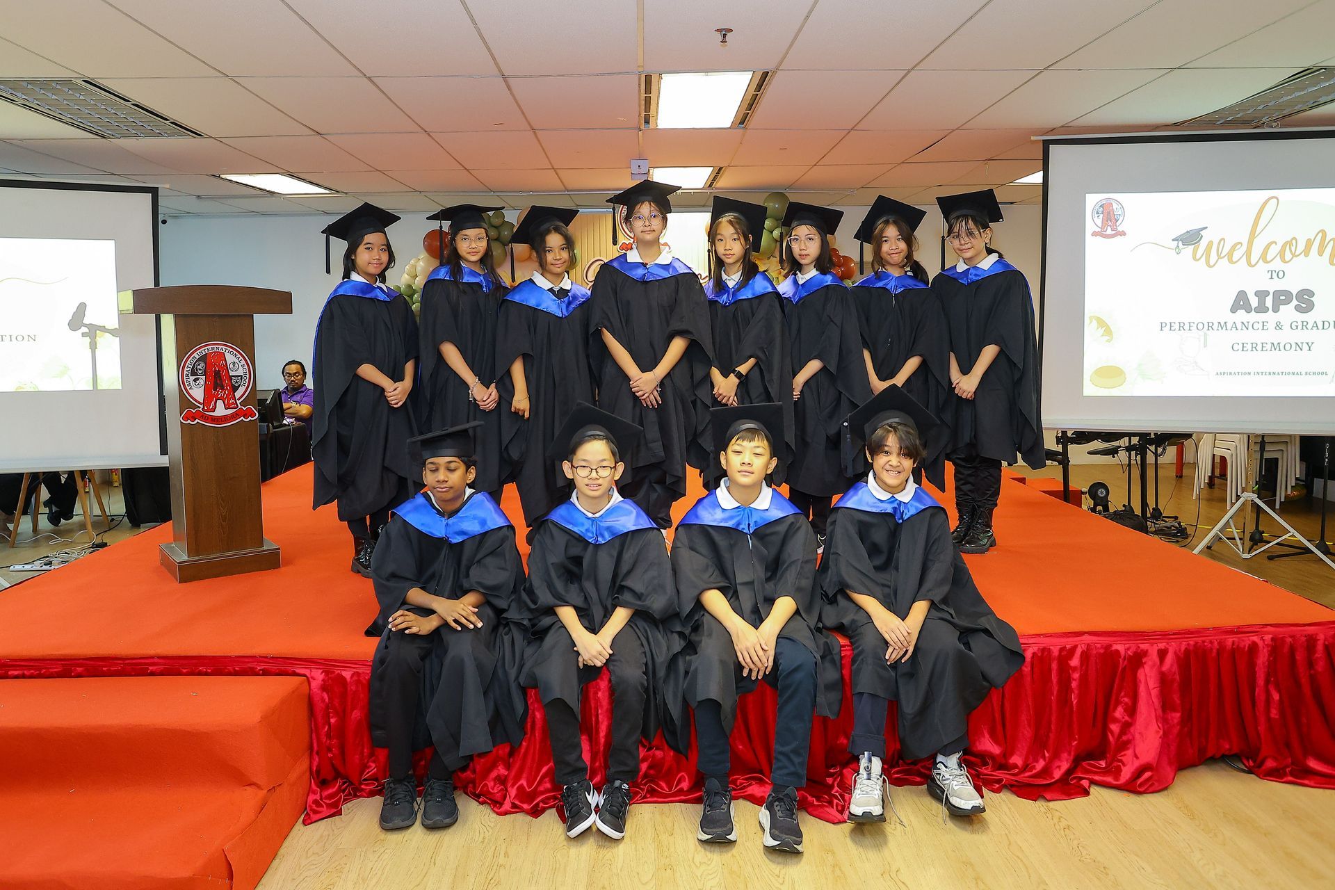 Students in graduation gowns pose on a stage with red skirting in a room with screens displaying welcome text.