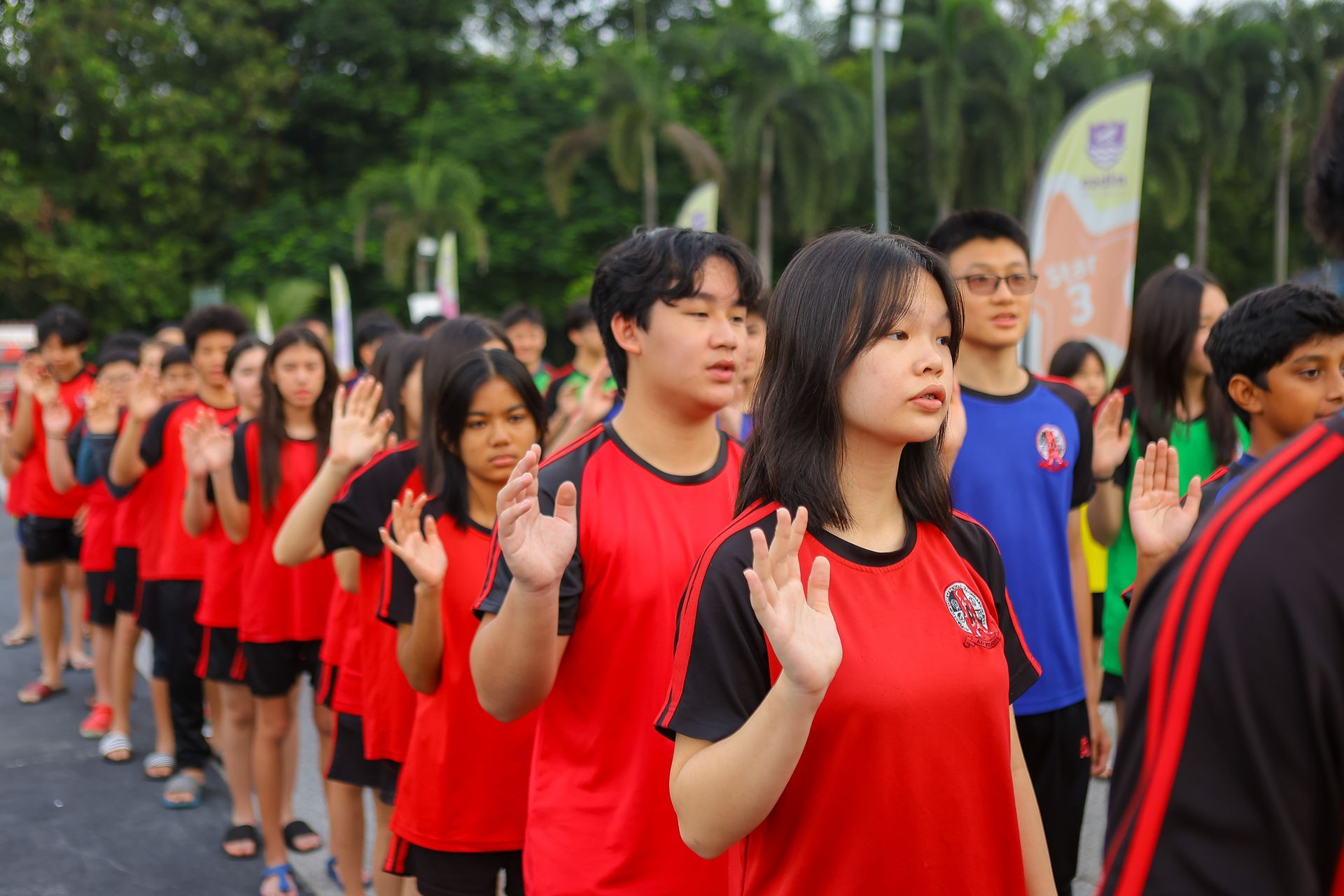 A group of students in red and blue uniforms stands in a line outdoors with their right hands raised.
