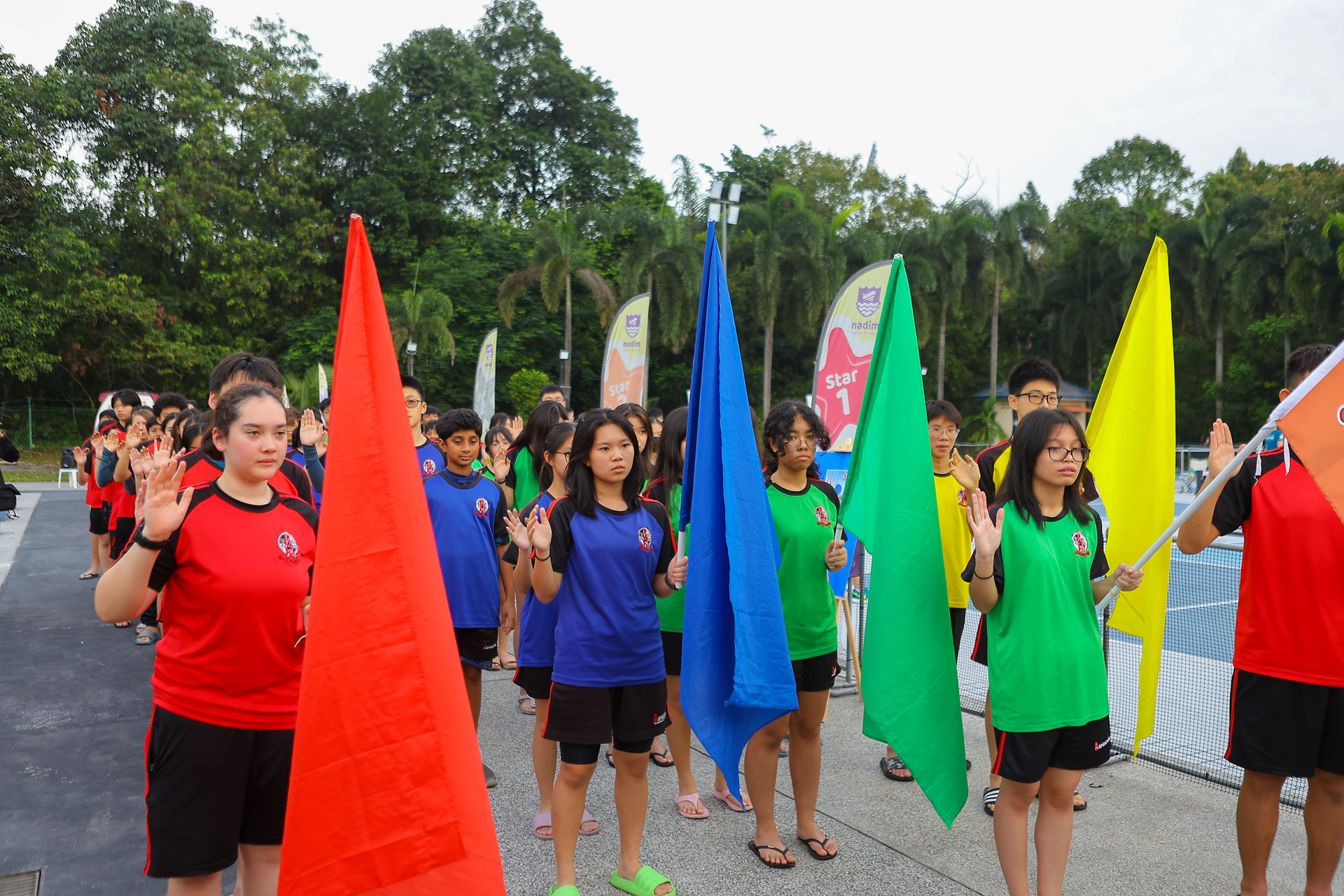 A group of students stand in an outdoor athletic area, holding colored flags and raising their right hands in an oath.