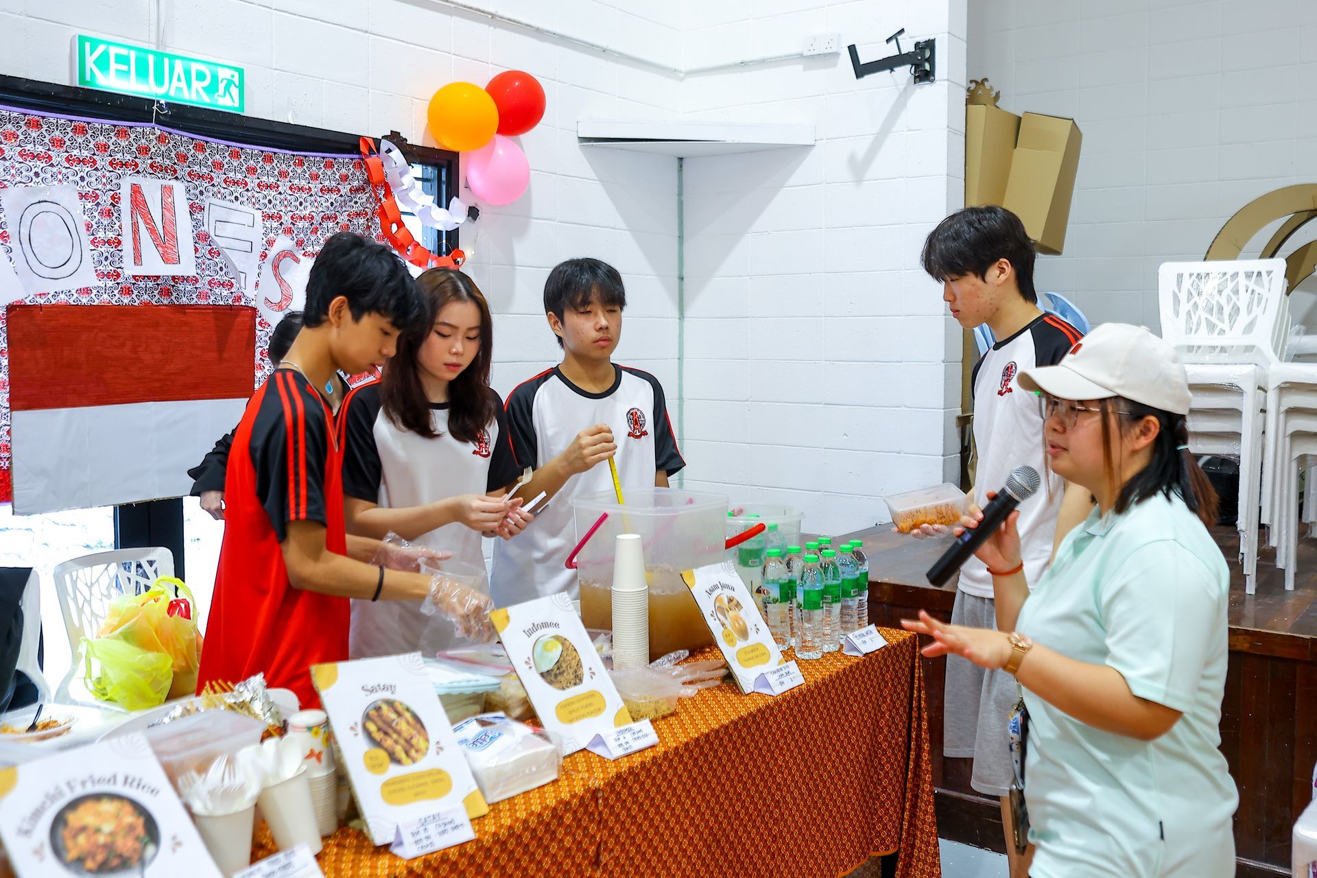Students stand behind a table with food and drinks at a booth, while a person with a microphone speaks to them.