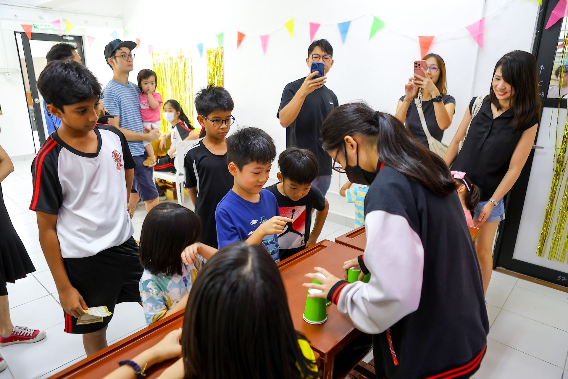 A group of people observe a person in a black jacket demonstrating a science activity with green cups on a wooden table.
