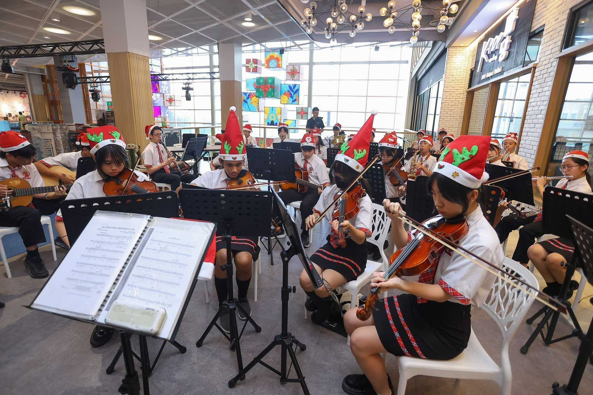 A student orchestra performs in a brightly lit atrium, musicians wearing Santa hats while playing violins.