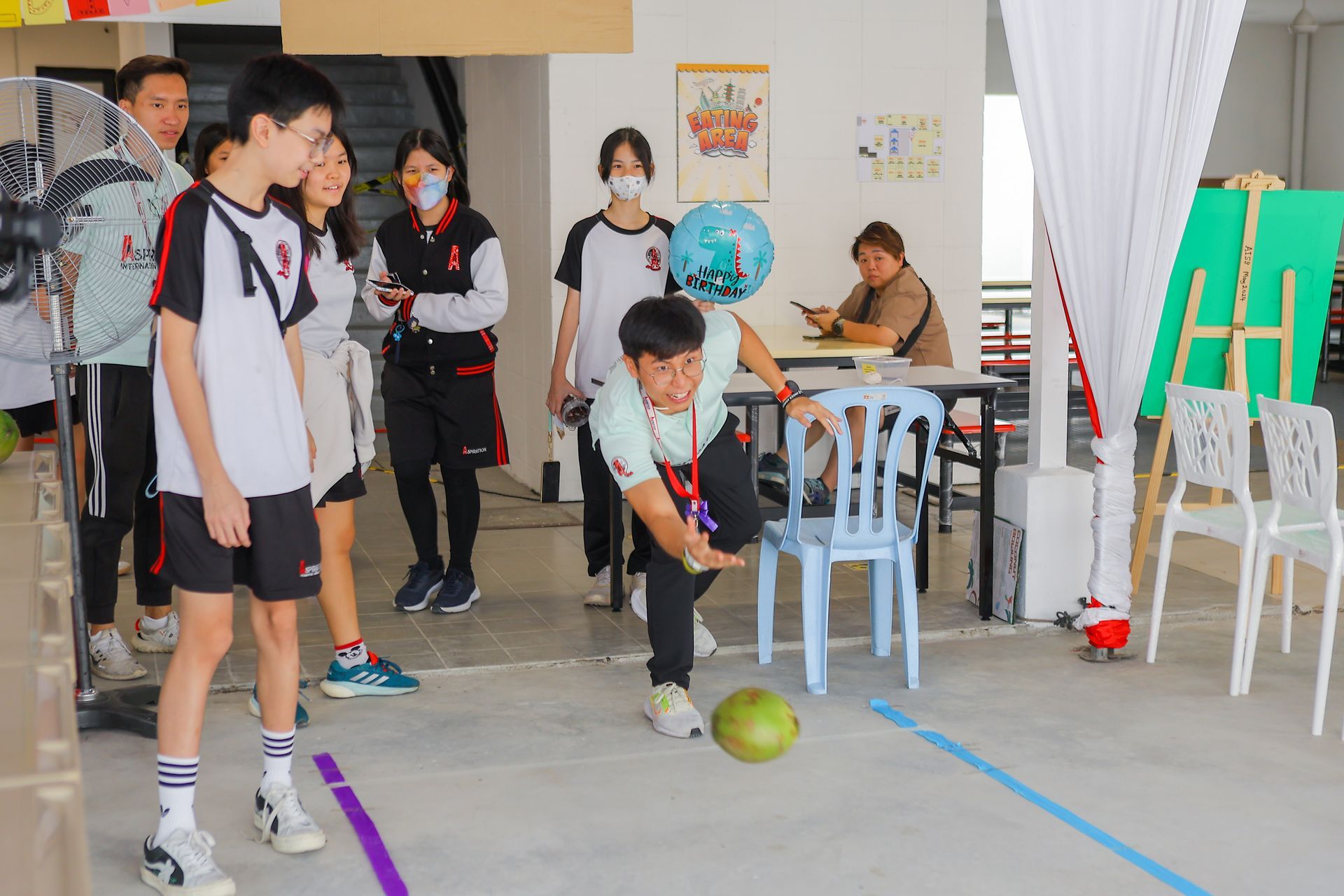 A person in a light green shirt rolls a coconut across the floor toward a blue ball while others watch in a room.