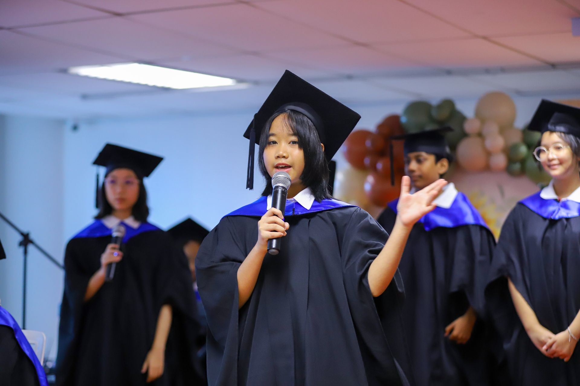 A person in a graduation gown and cap speaks into a microphone at an event, with classmates standing in the background.