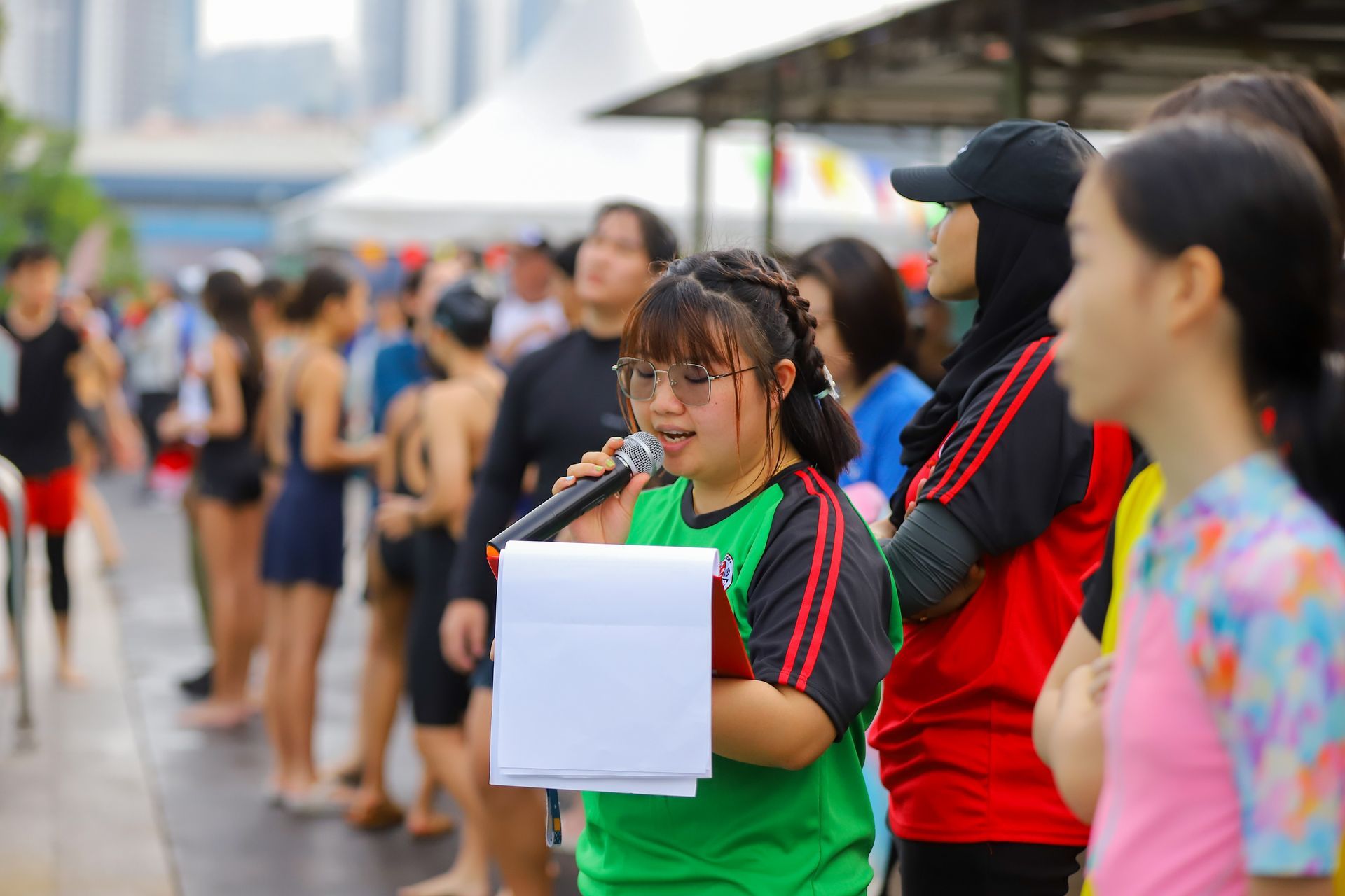 A person in a green shirt speaks into a microphone while holding a clipboard at an outdoor event near a pool.
