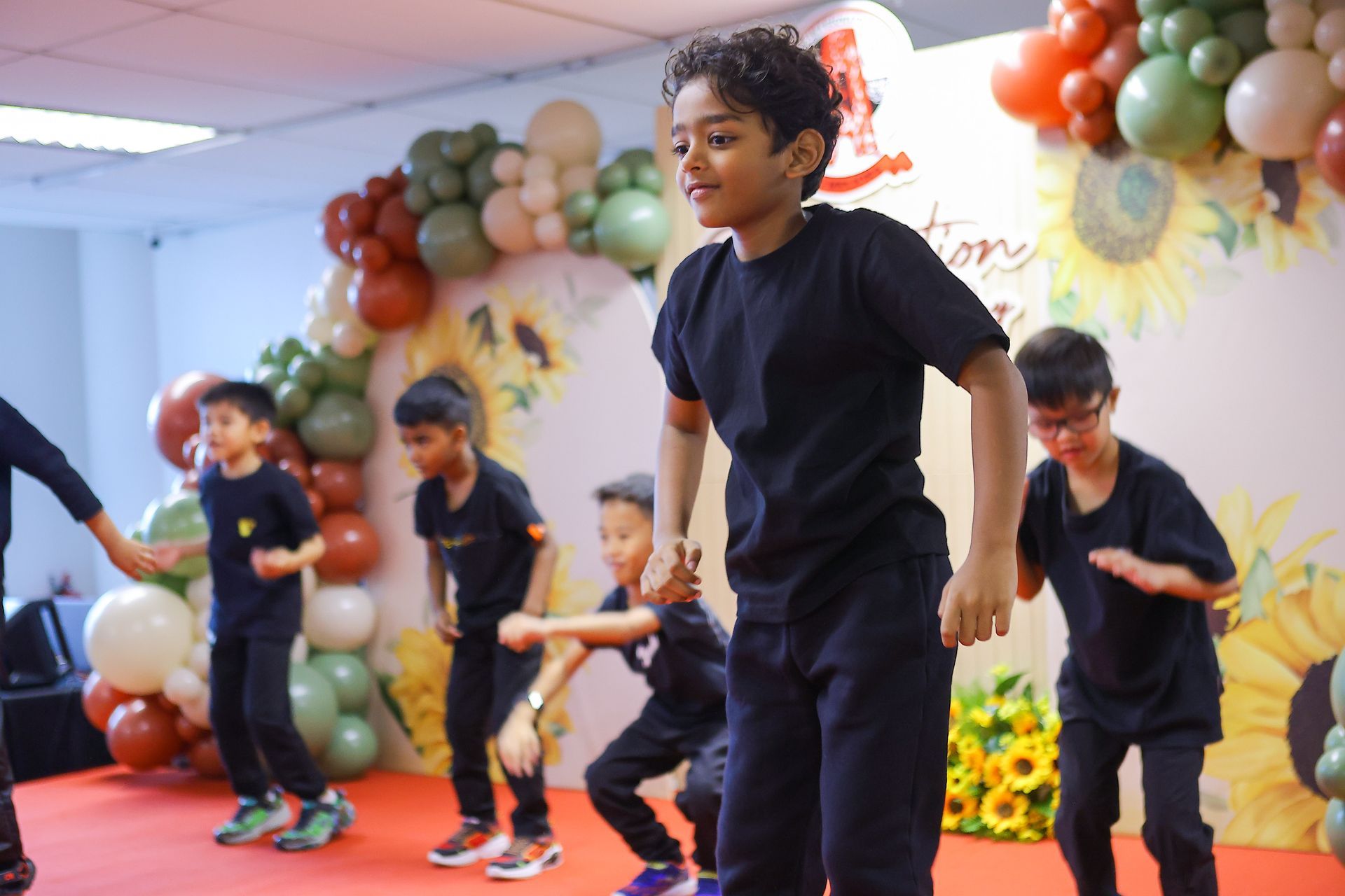 Five children in matching black shirts perform a choreographed dance on a stage decorated with balloons and sunflowers.