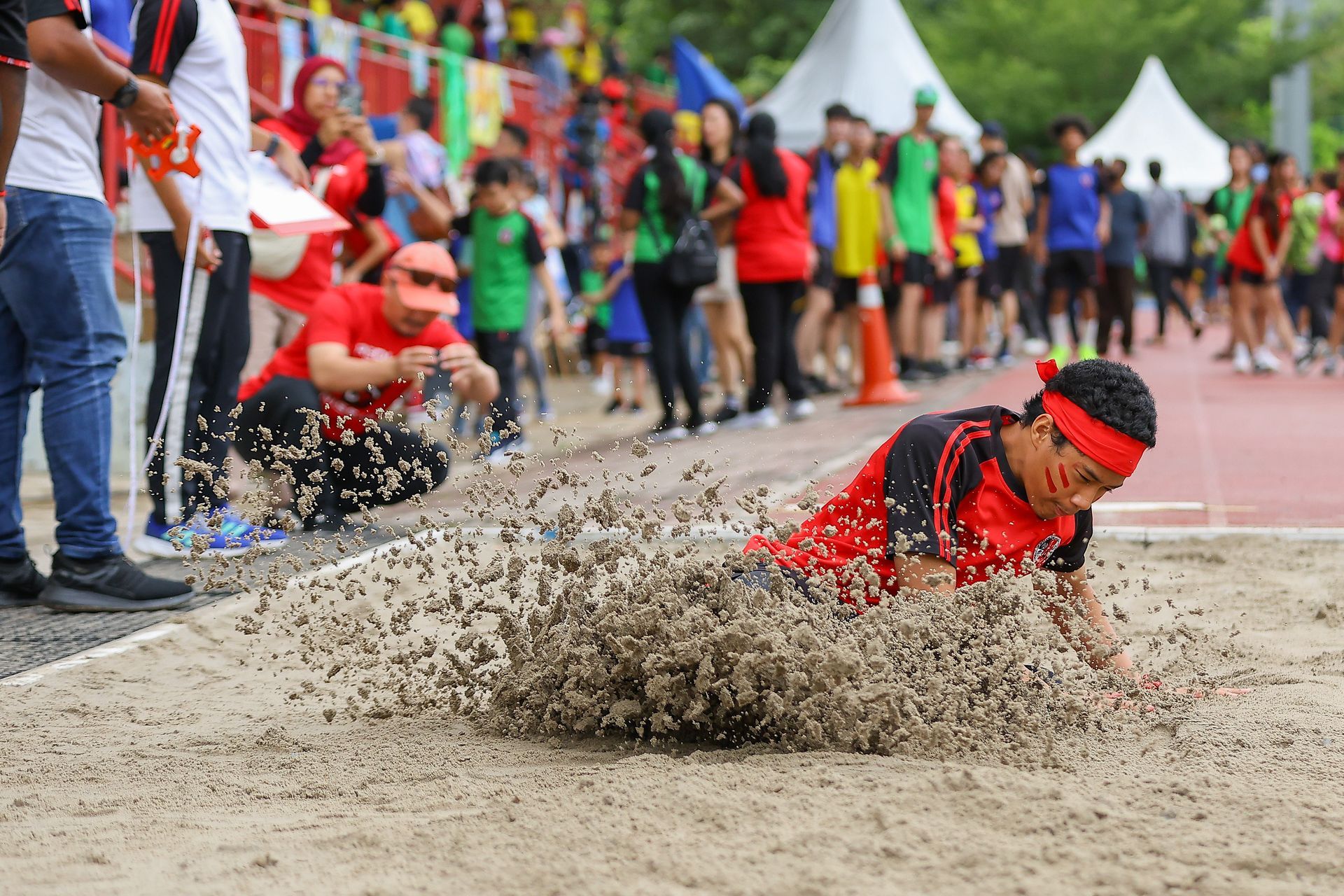 A long jumper lands in the sand pit during an outdoor track and field competition, with spectators watching in the back.